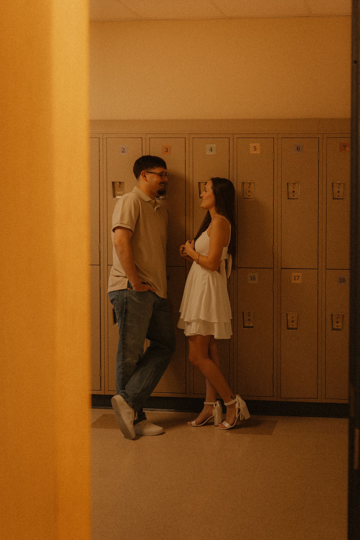 Couple standing by school lockers in warm light, woman in white dress and man in casual outfit facing each other.
