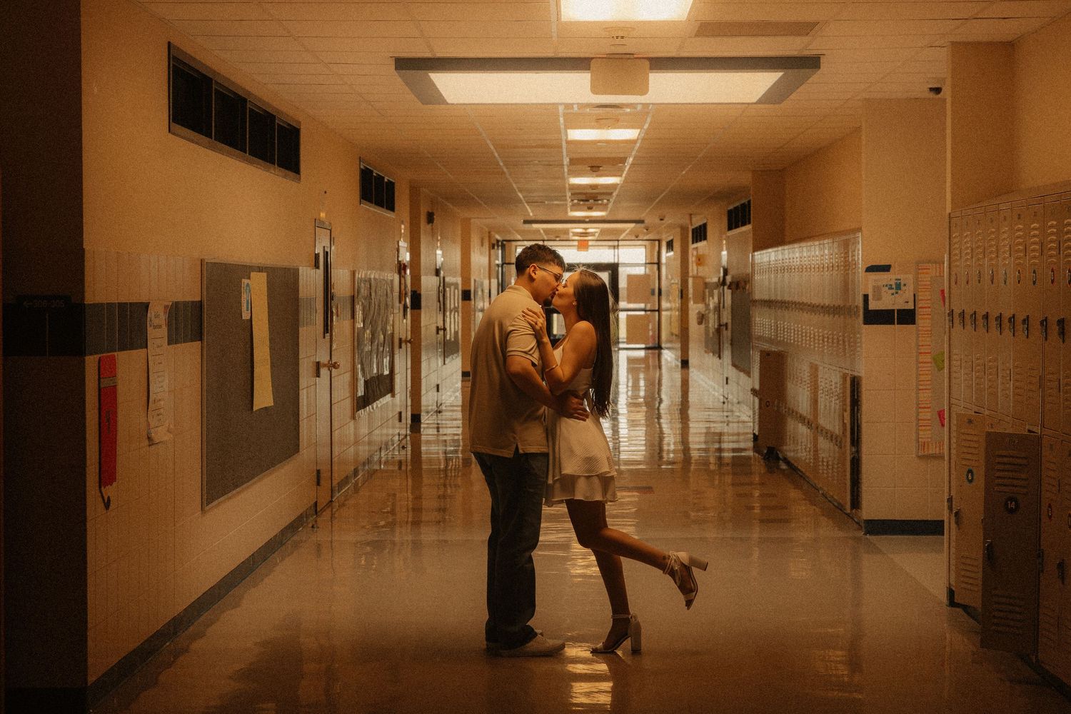 Couple sharing a romantic kiss in a warmly lit school hallway with lockers lining the walls.