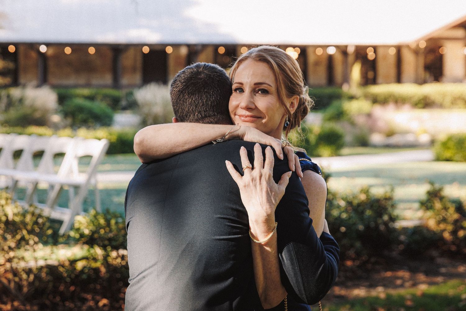 Emotional bride hugging groom outdoors showing engagement ring with wedding venue in background.