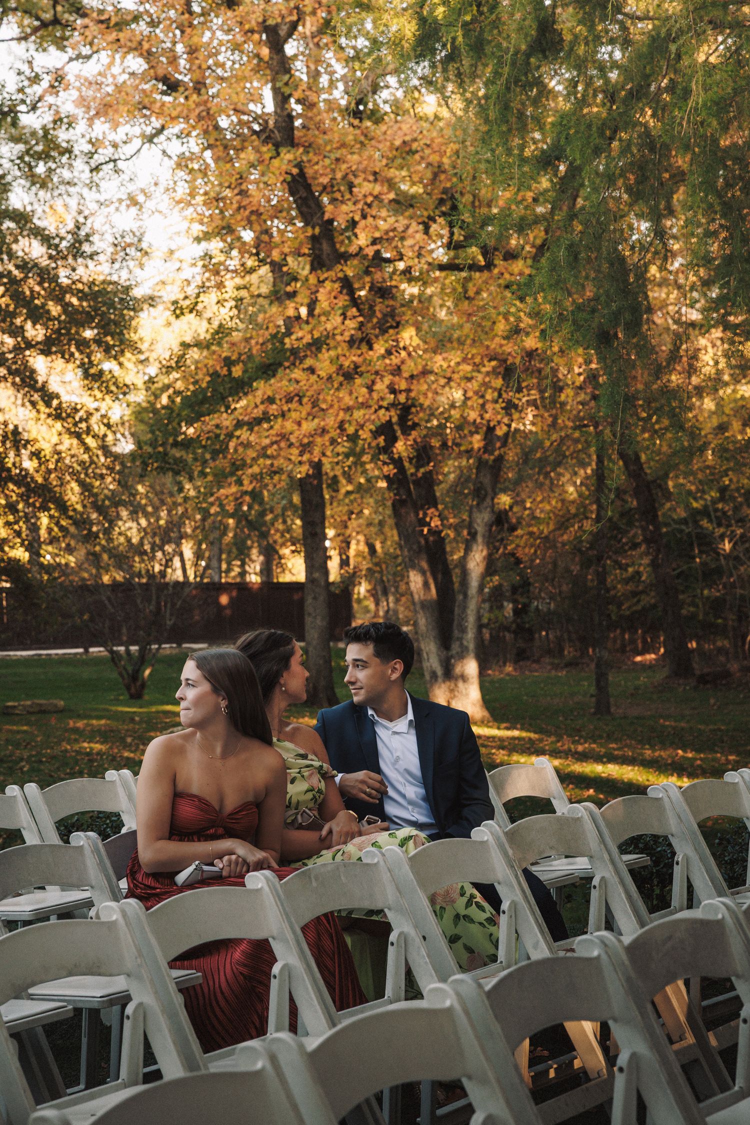 Wedding guests in formal attire seated in white chairs at outdoor autumn ceremony.