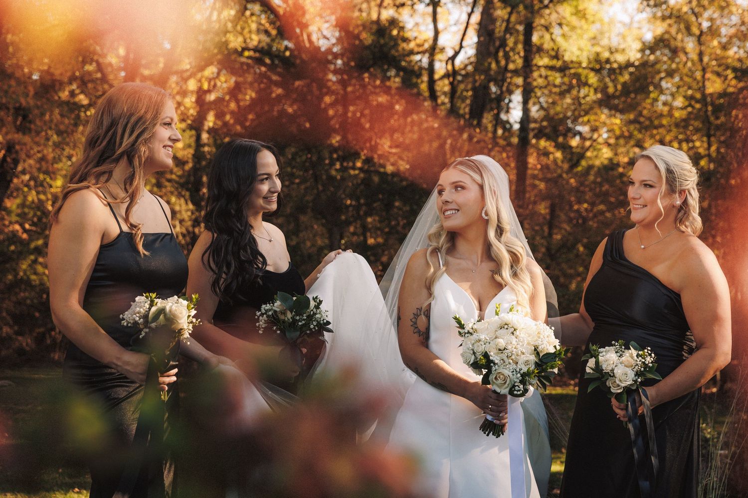 Bride in white dress with veil and three bridesmaids in black gowns in autumn light.