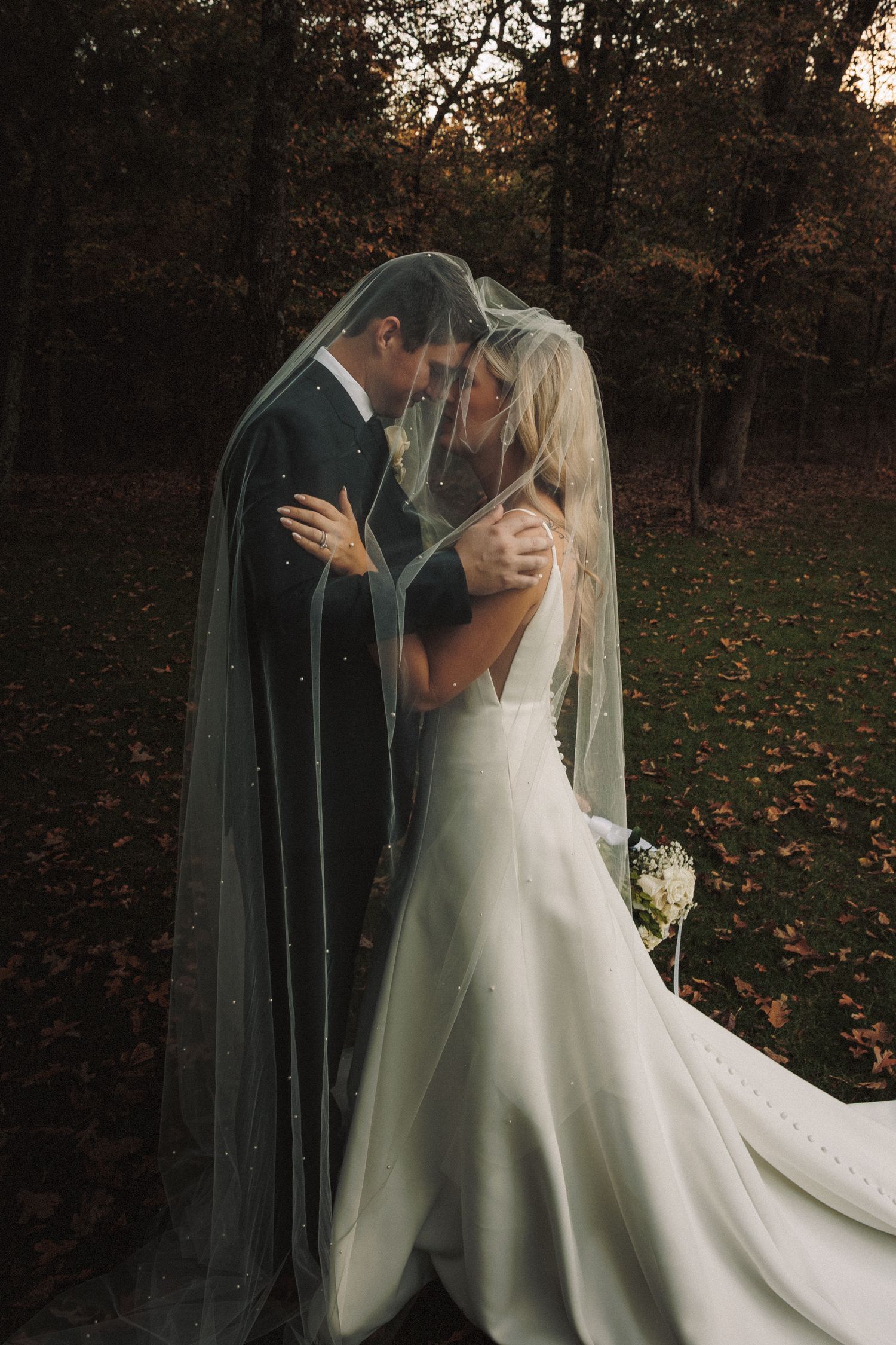Bride and groom share an intimate moment under a pearl-adorned wedding veil surrounded by autumn foliage.