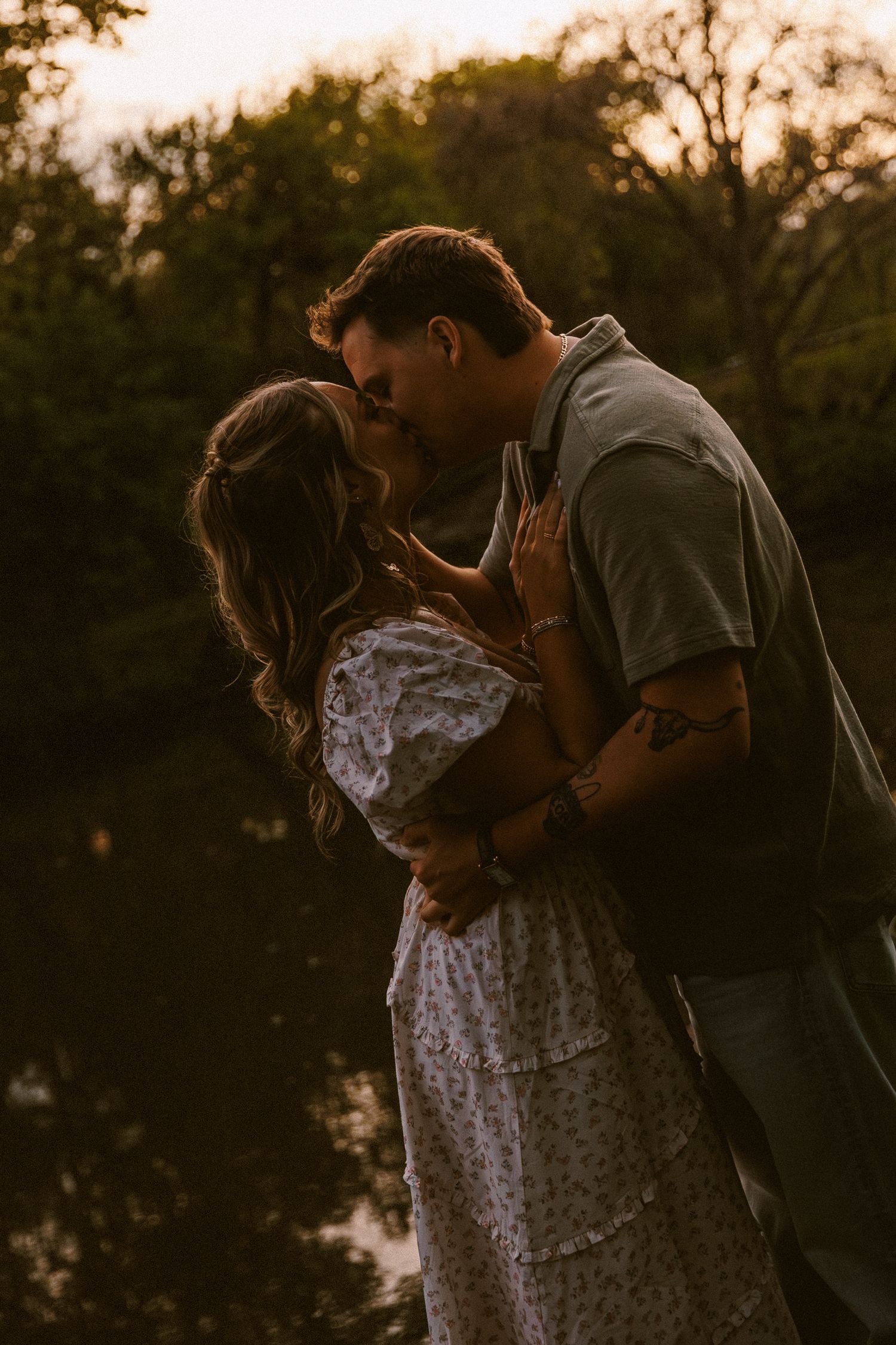 Couple sharing a kiss near a pond at sunset, surrounded by dark trees.