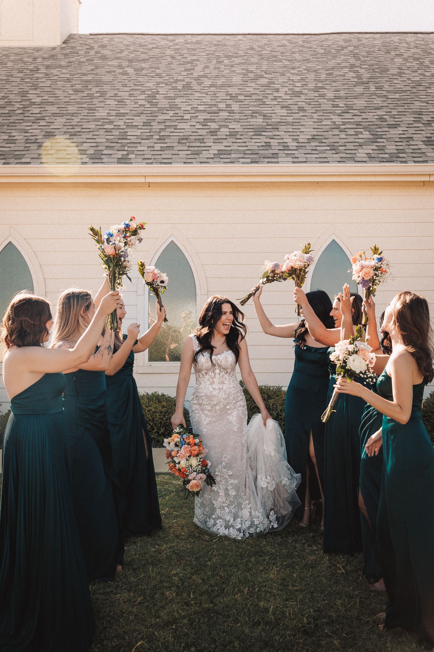 Bride in lace gown walks through arch of raised bouquets held by bridesmaids in emerald green dresses outside a chapel.
