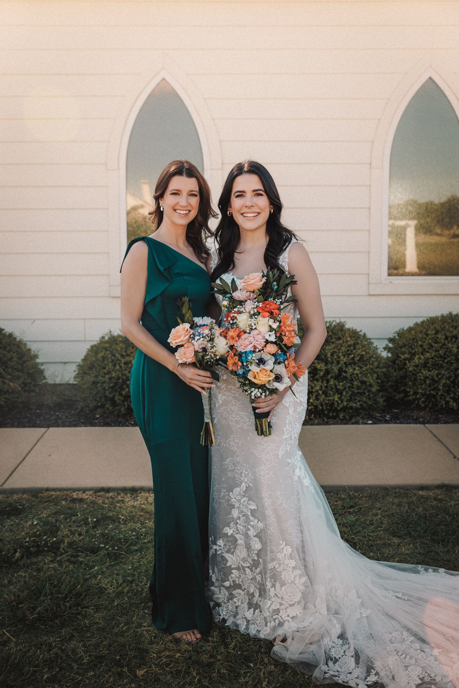 Bride in lace gown with bridesmaid in emerald dress holding colorful bouquets outside chapel.