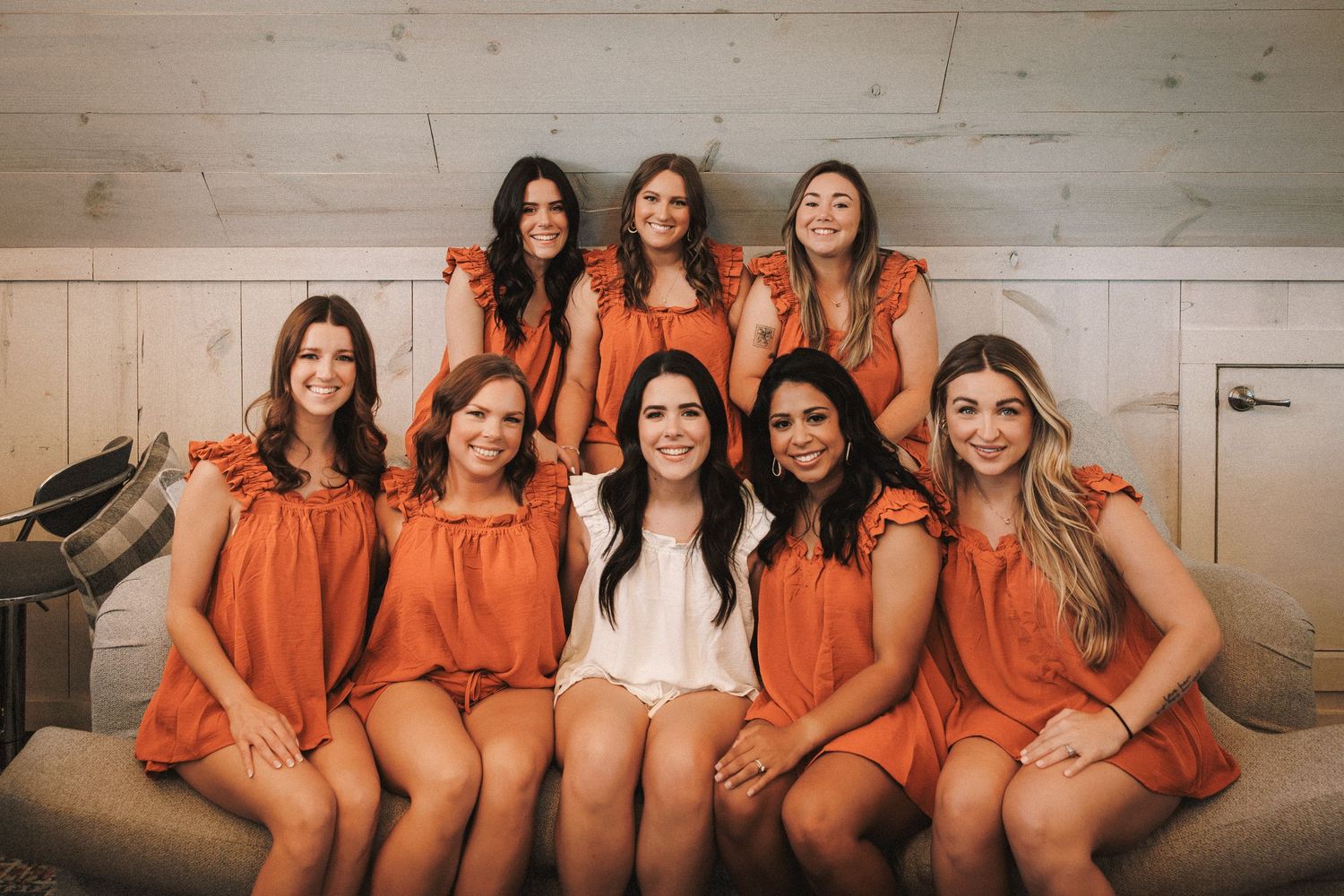 Group of bridesmaids in orange dresses posing with bride in white on a couch.