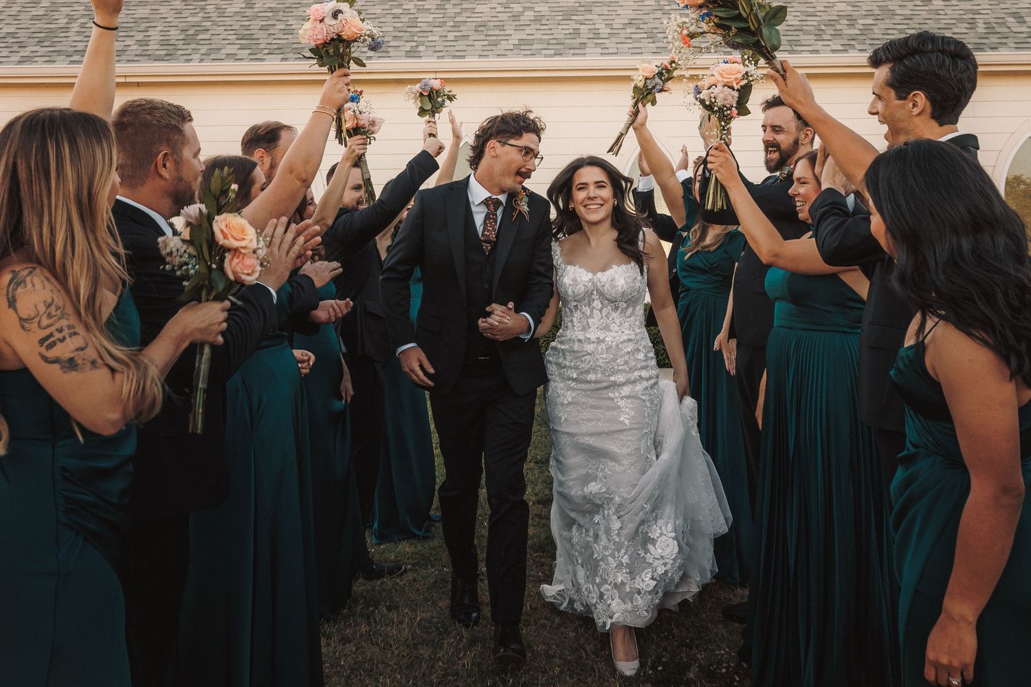 Newlyweds exit under raised bouquets as wedding party in emerald and black celebrates outside white chapel.