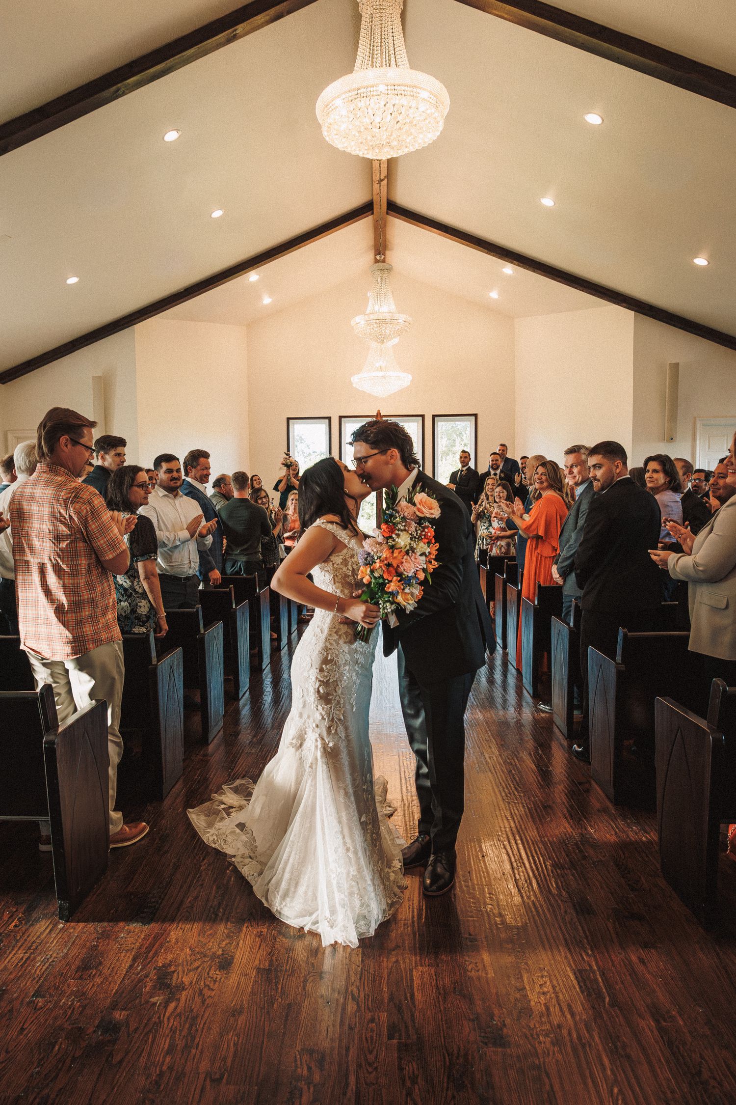 Newlyweds share first kiss in chapel aisle surrounded by standing guests and chandelier overhead.