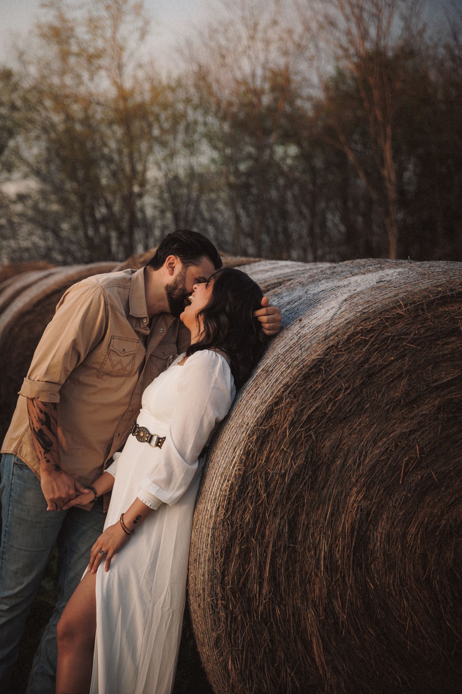 Couple kissing beside a large round hay bale on a farm at golden hour.