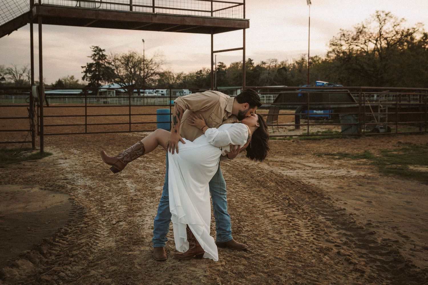 Romantic couple sharing passionate dip kiss at sunset in sandy ranch arena.