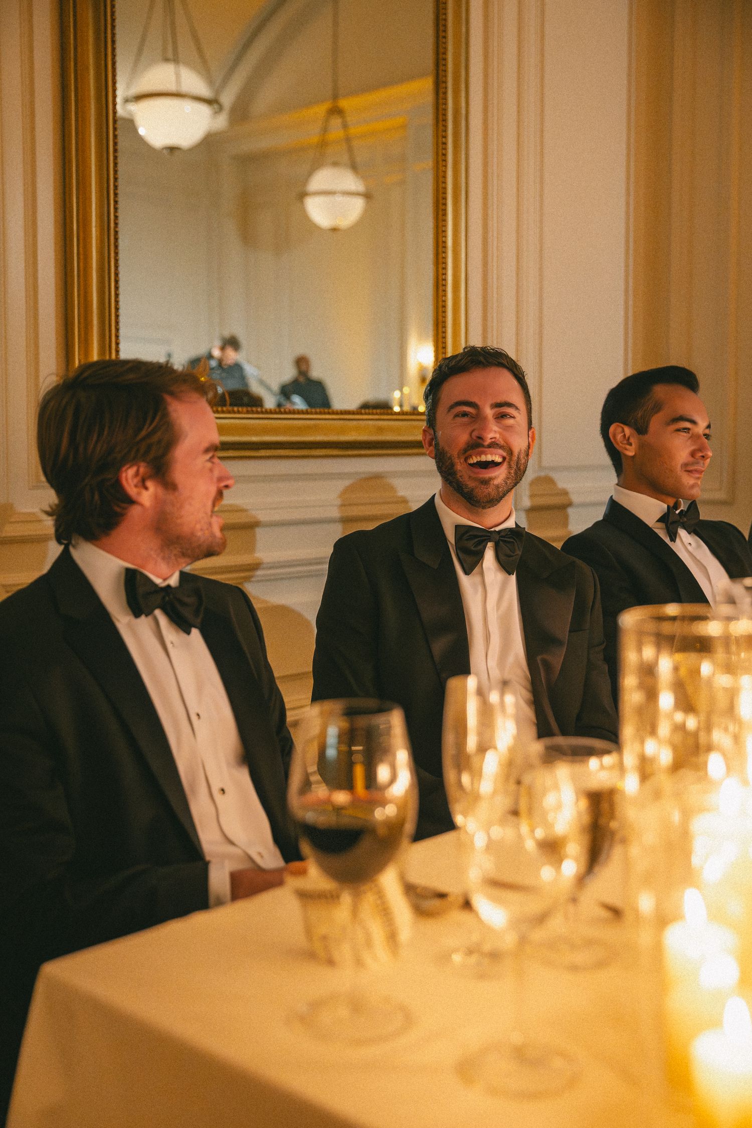 Three men in black tuxedos laugh joyfully at a formal wedding reception dinner table.