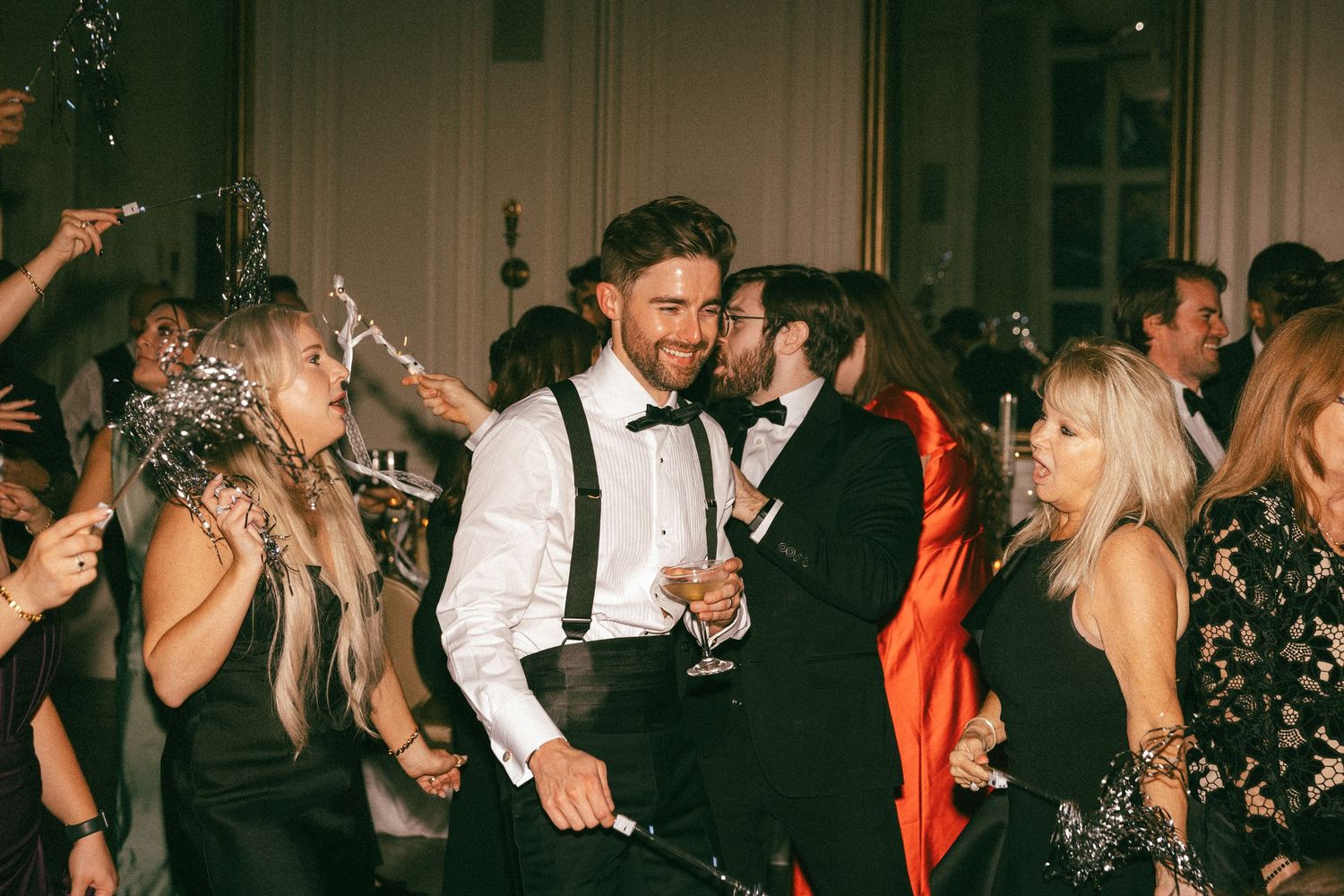 Groom in suspenders celebrating with champagne surrounded by guests at wedding party.