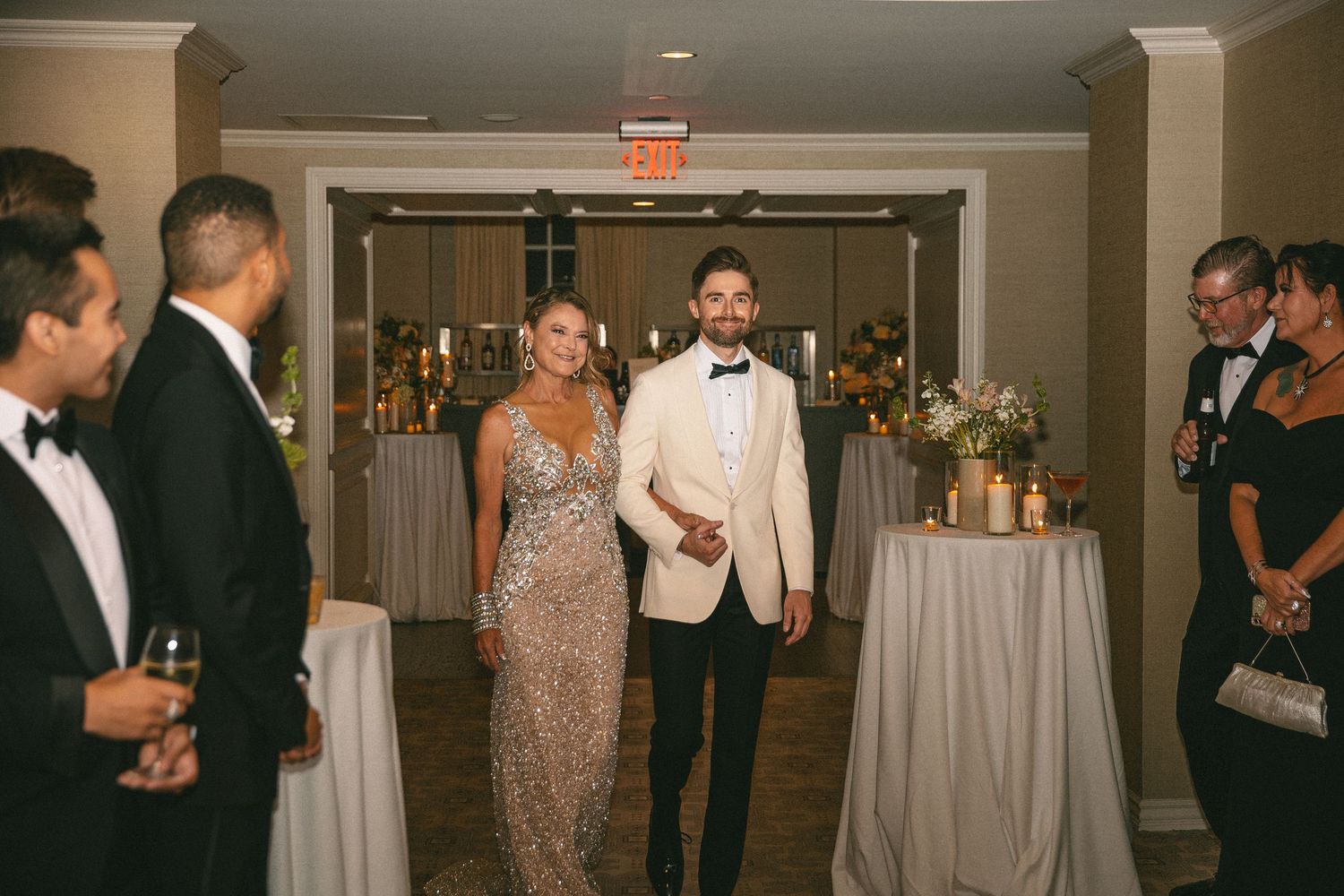 Groom in white tuxedo walks with a woman in a sequined gown during wedding reception entrance.