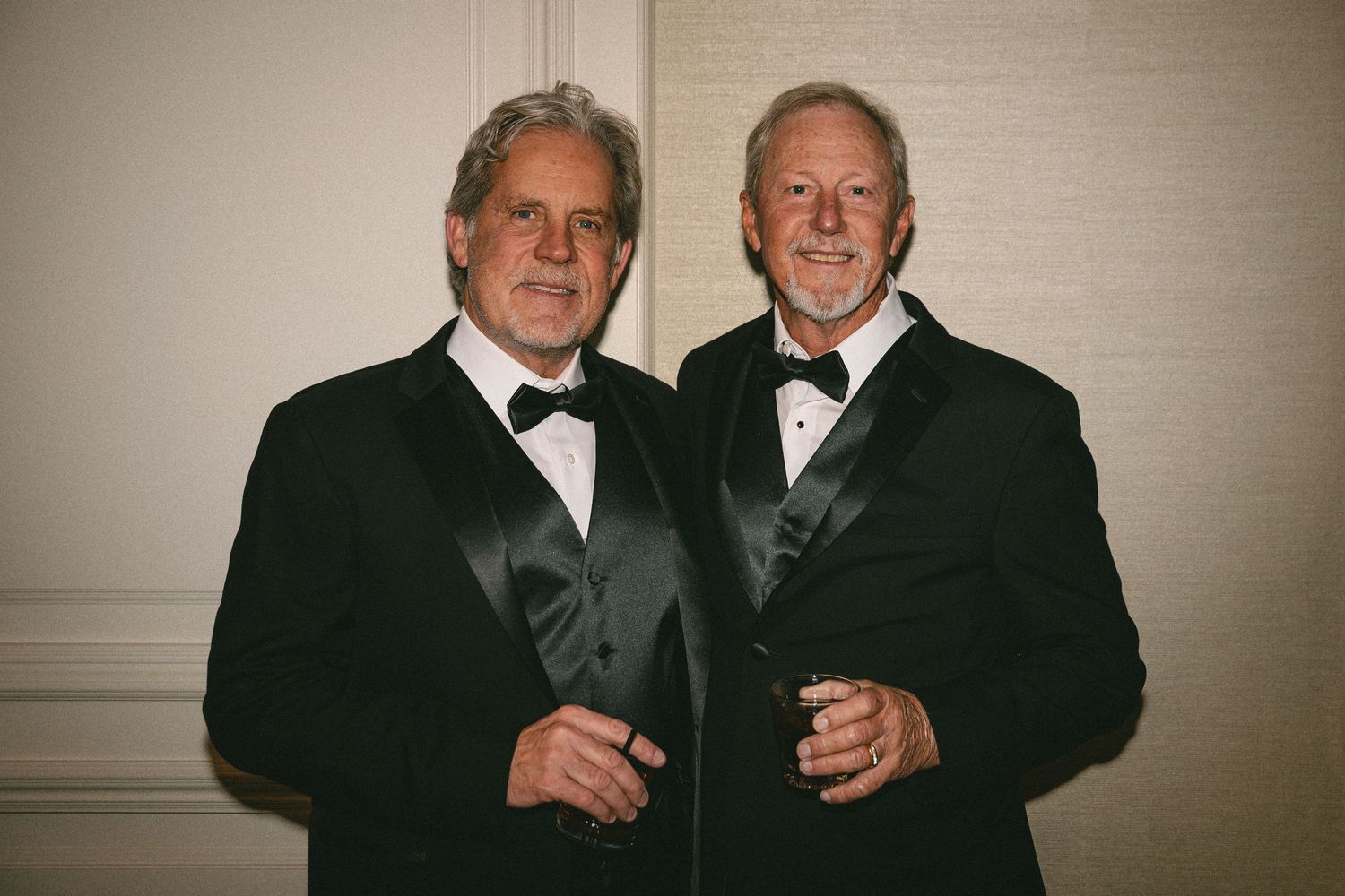 Two men in classic black tuxedos smile together at a formal wedding reception.