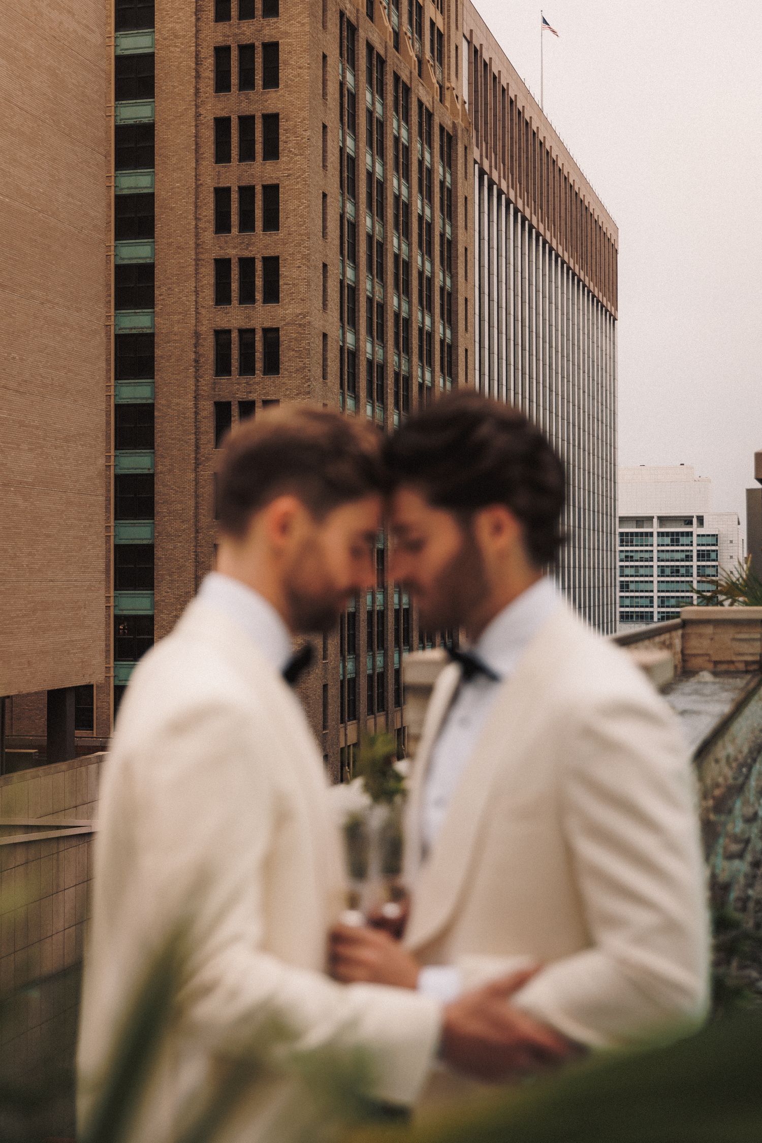 Two grooms in white tuxedos sharing an intimate moment on a rooftop with city buildings in background.