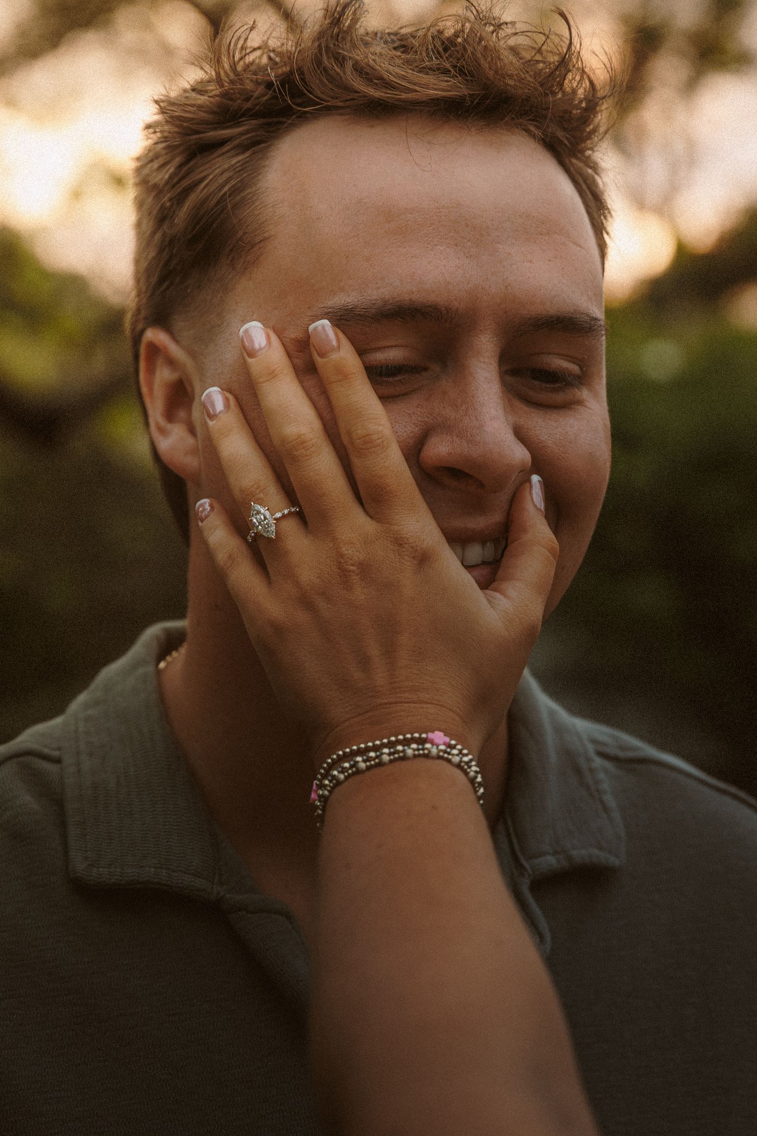 Person laughing with hand on face, wearing diamond ring and pink beaded bracelet, outdoors at golden hour.