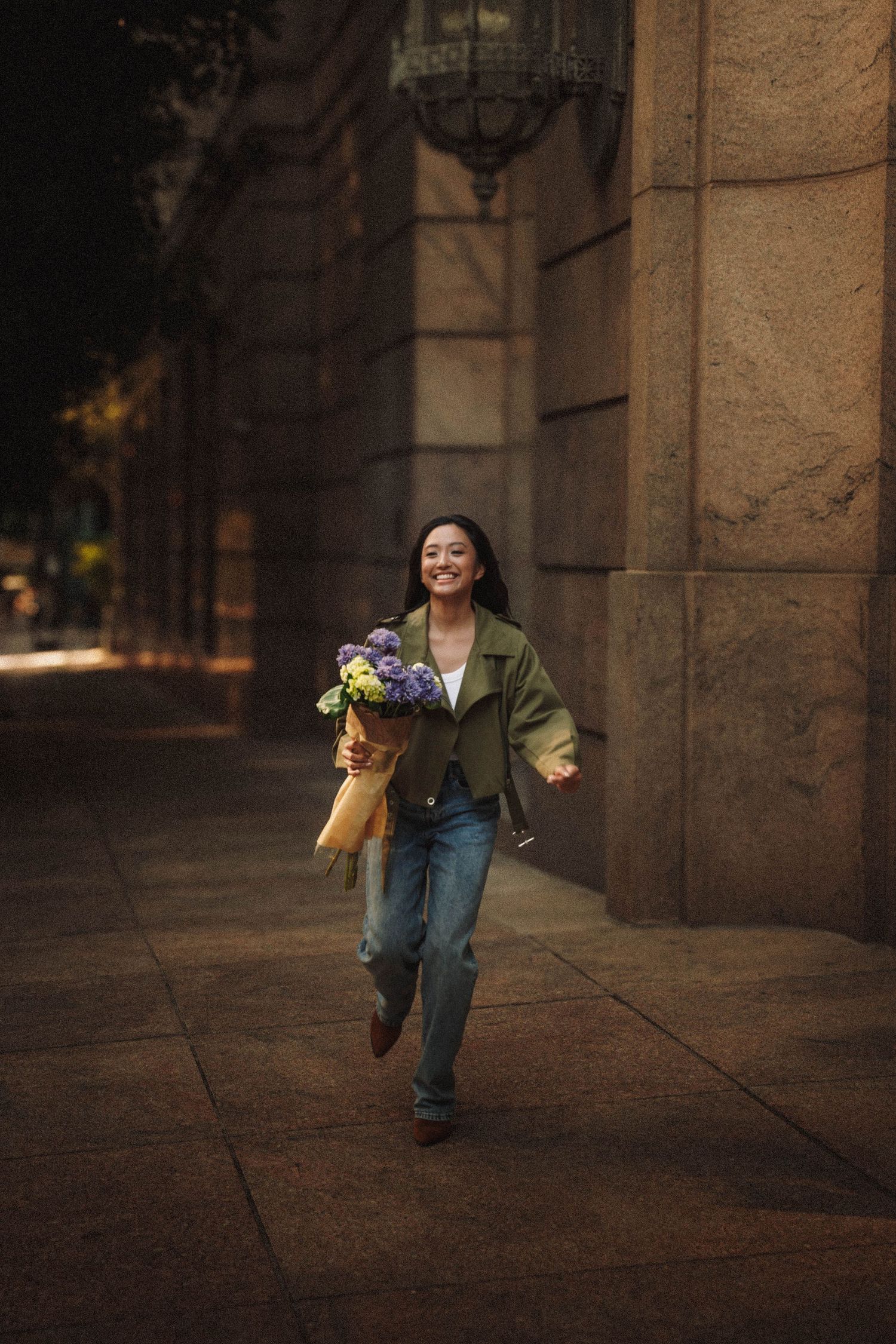 A person runs joyfully through a stone archway while holding a bright bouquet of flowers and smiling at dusk.