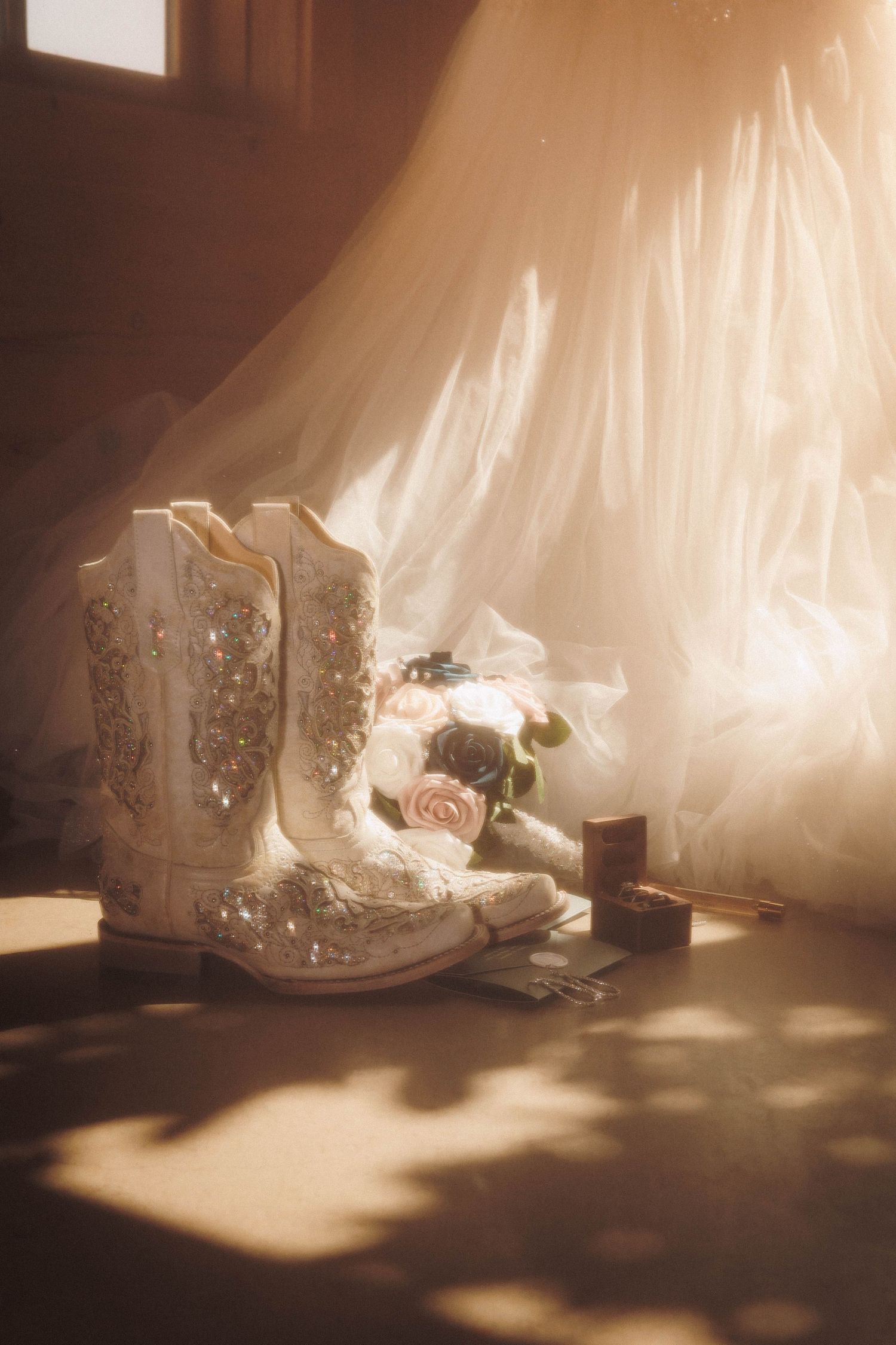 A pair of white embellished cowboy boots with roses and a veil in soft sepia lighting create a romantic wedding setting.