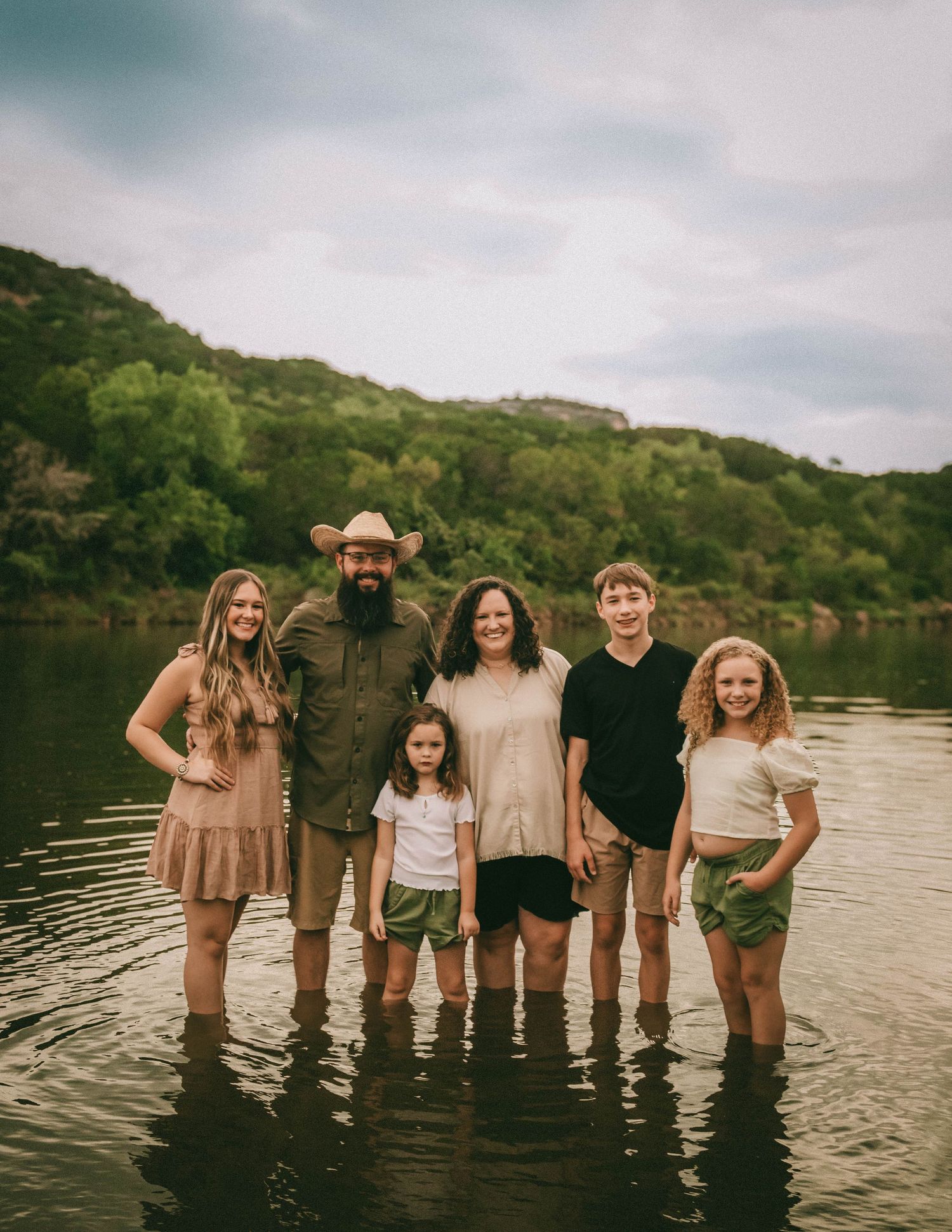 A family group stands together in a lake with hills in the background at sunset creating a serene outdoor portrait.