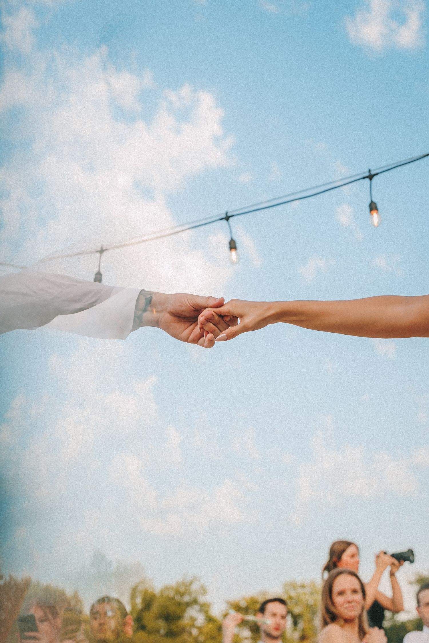 Artistic shot of hands reaching towards each other against a blue sky with string lights.