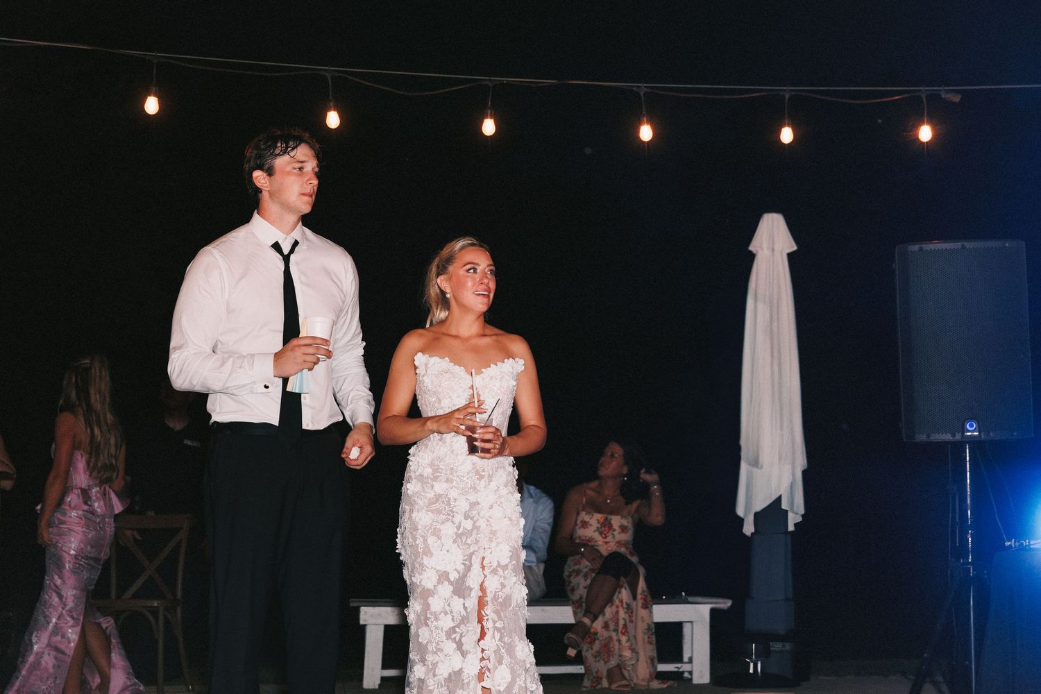 Wedding guests dancing and celebrating on darkly lit dance floor with string lights overhead during evening reception.