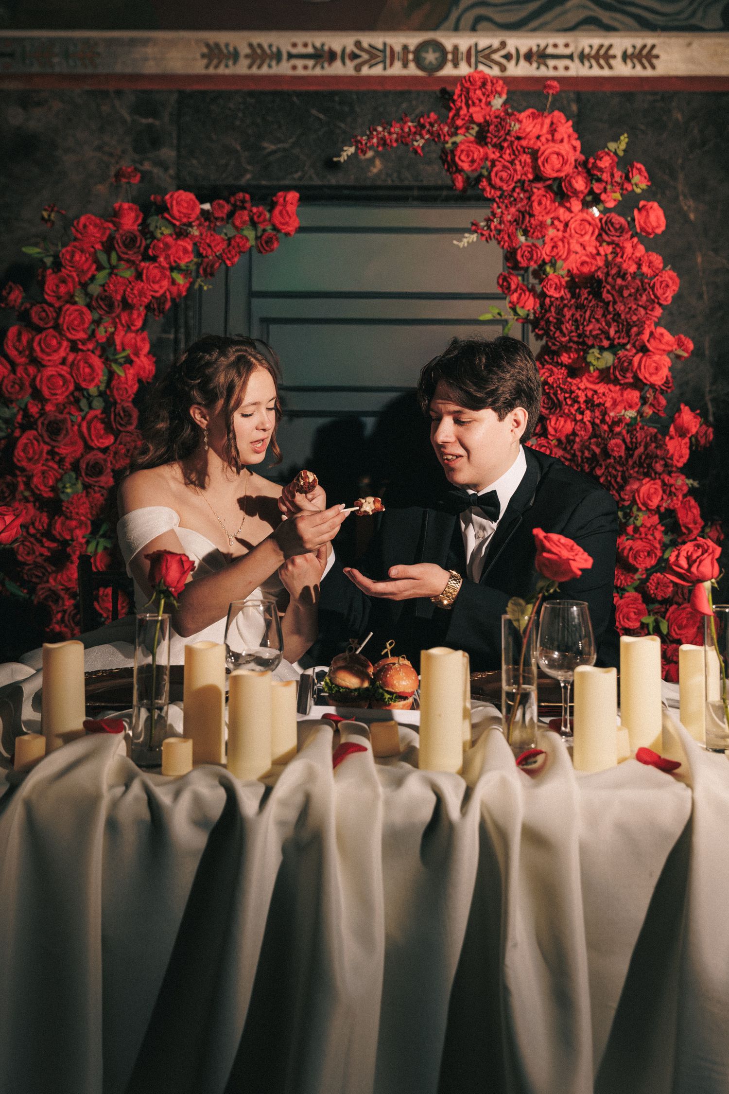Bride and groom share food at sweetheart table adorned with red roses and pillar candles.