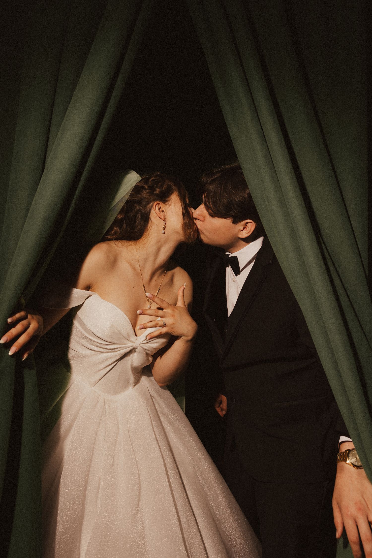 Bride and groom sharing romantic kiss between emerald green velvet curtains at wedding ceremony.