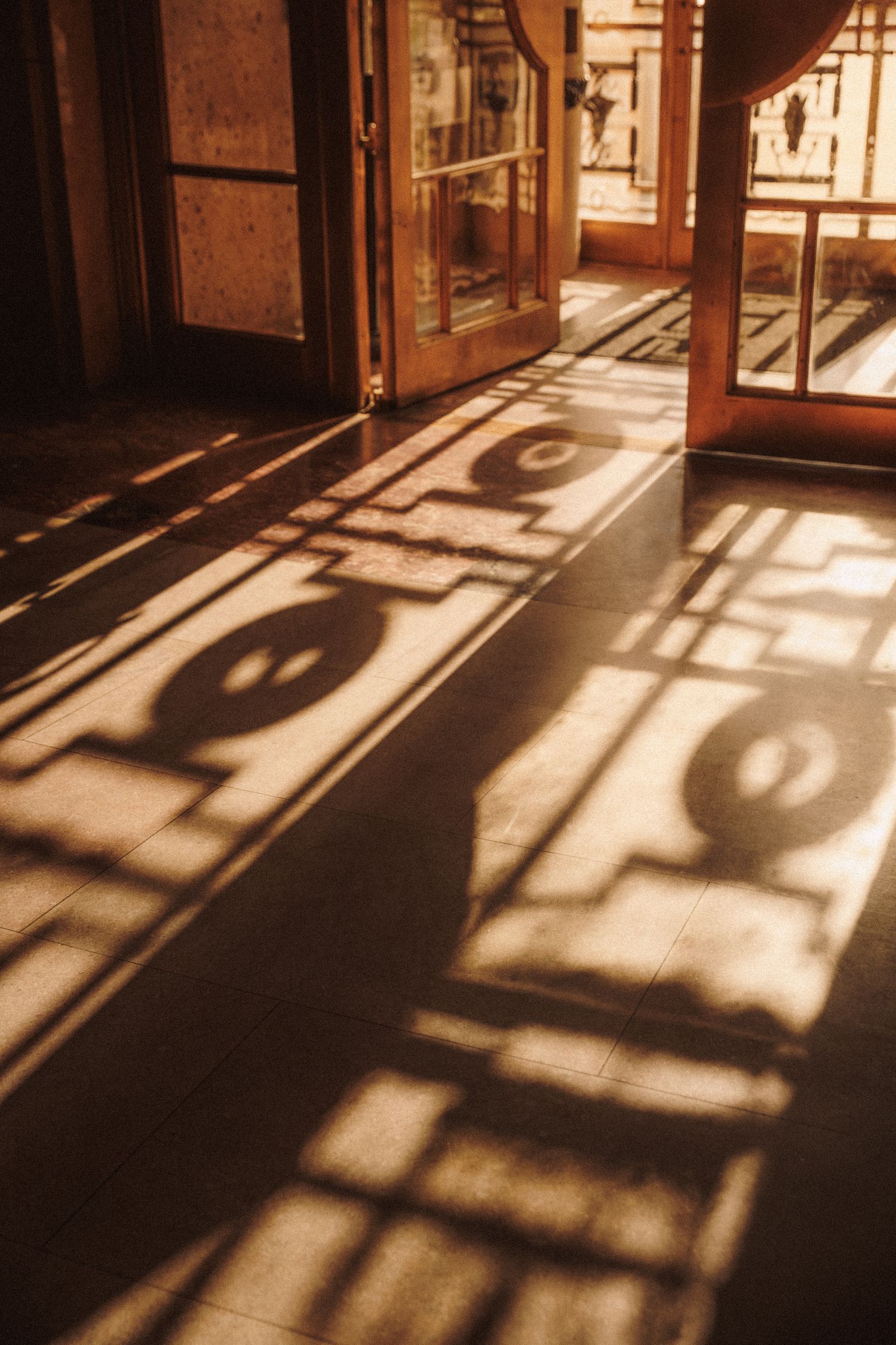 Warm golden sunlight casting ornate iron gate shadows on tiled floor through open wooden doors.