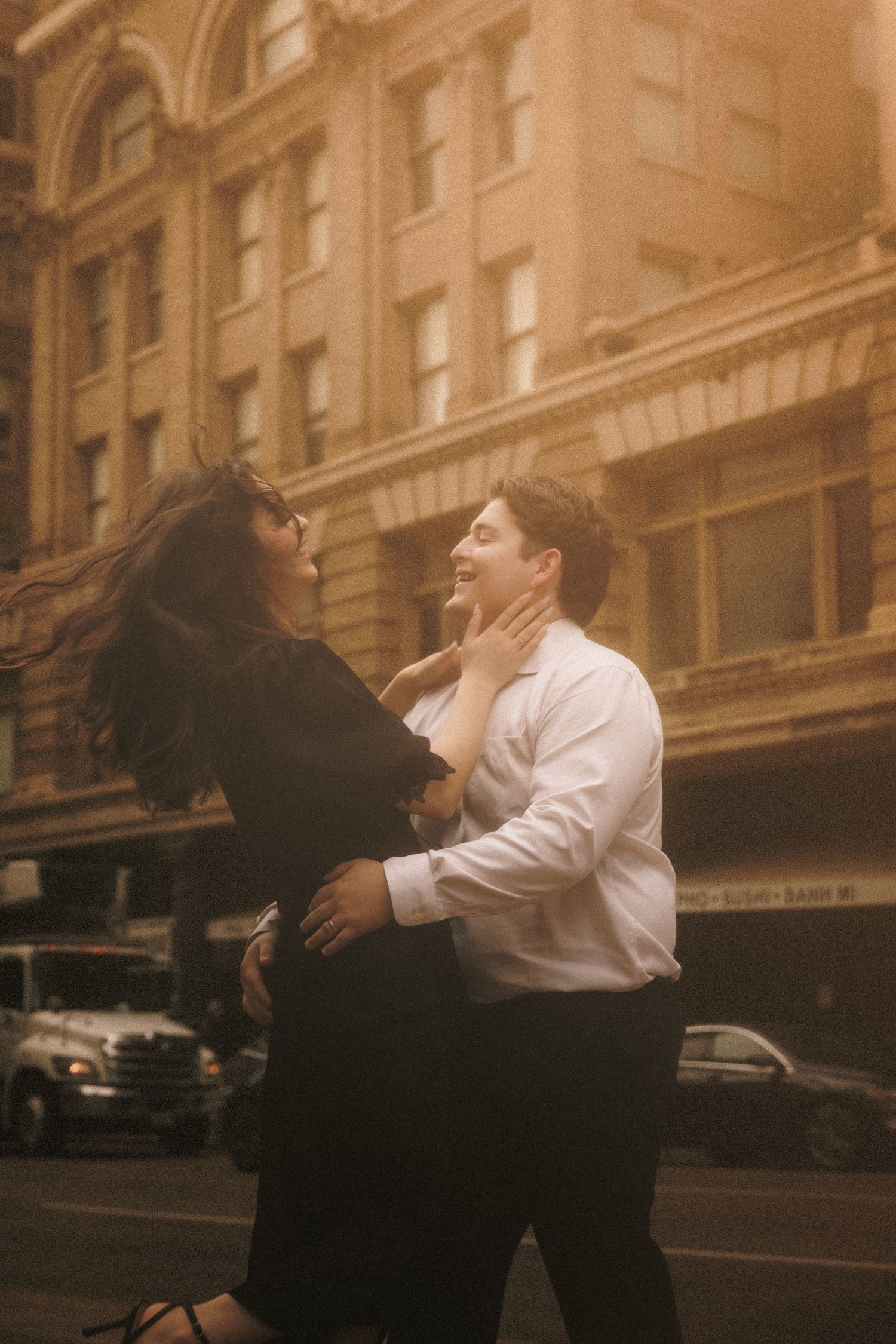 Couple dancing romantically on a city street at golden hour, woman in black dress touching man's face in urban setting.