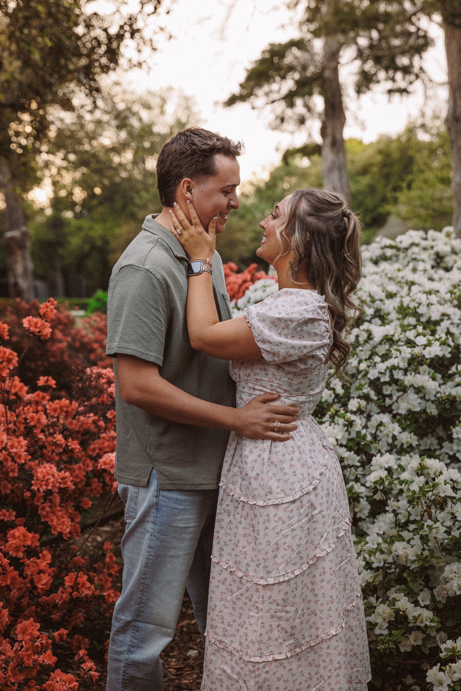 Couple embracing and smiling among red and white flowering bushes during golden hour in a garden.