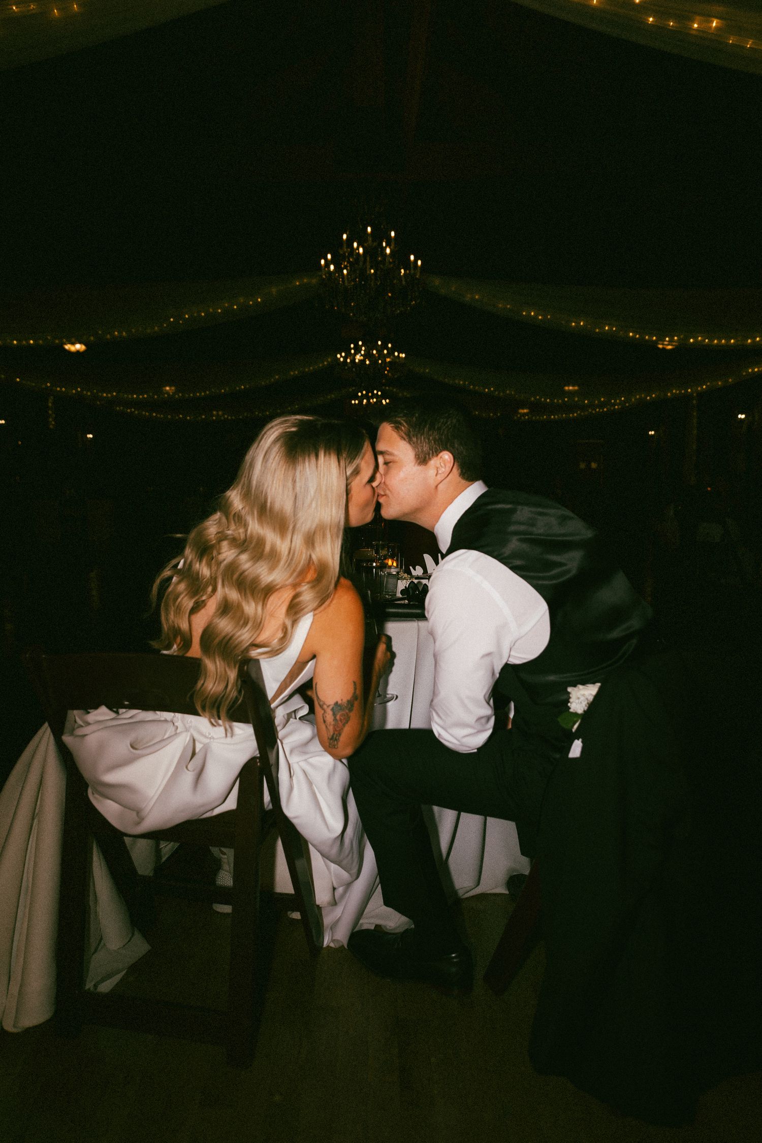 Bride and groom share a kiss at their reception table under warm string lights.