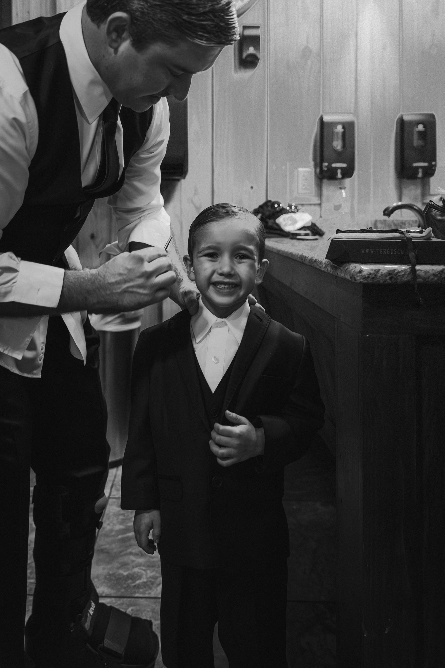 Black and white photo of groom helping smiling young ring bearer with suit in bathroom mirror.