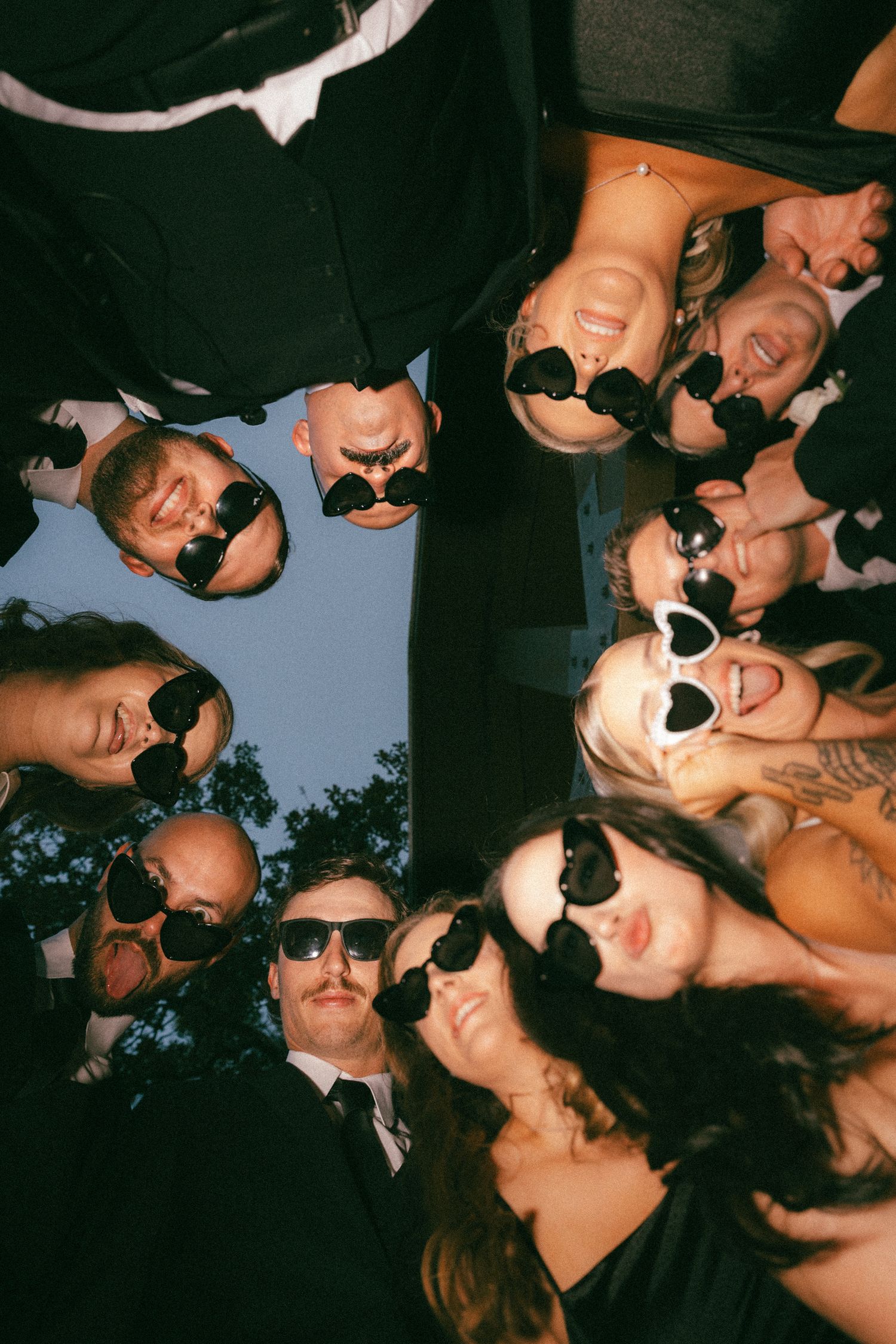 Wedding party in sunglasses forms a fun circle huddle photographed from below against a blue sky.