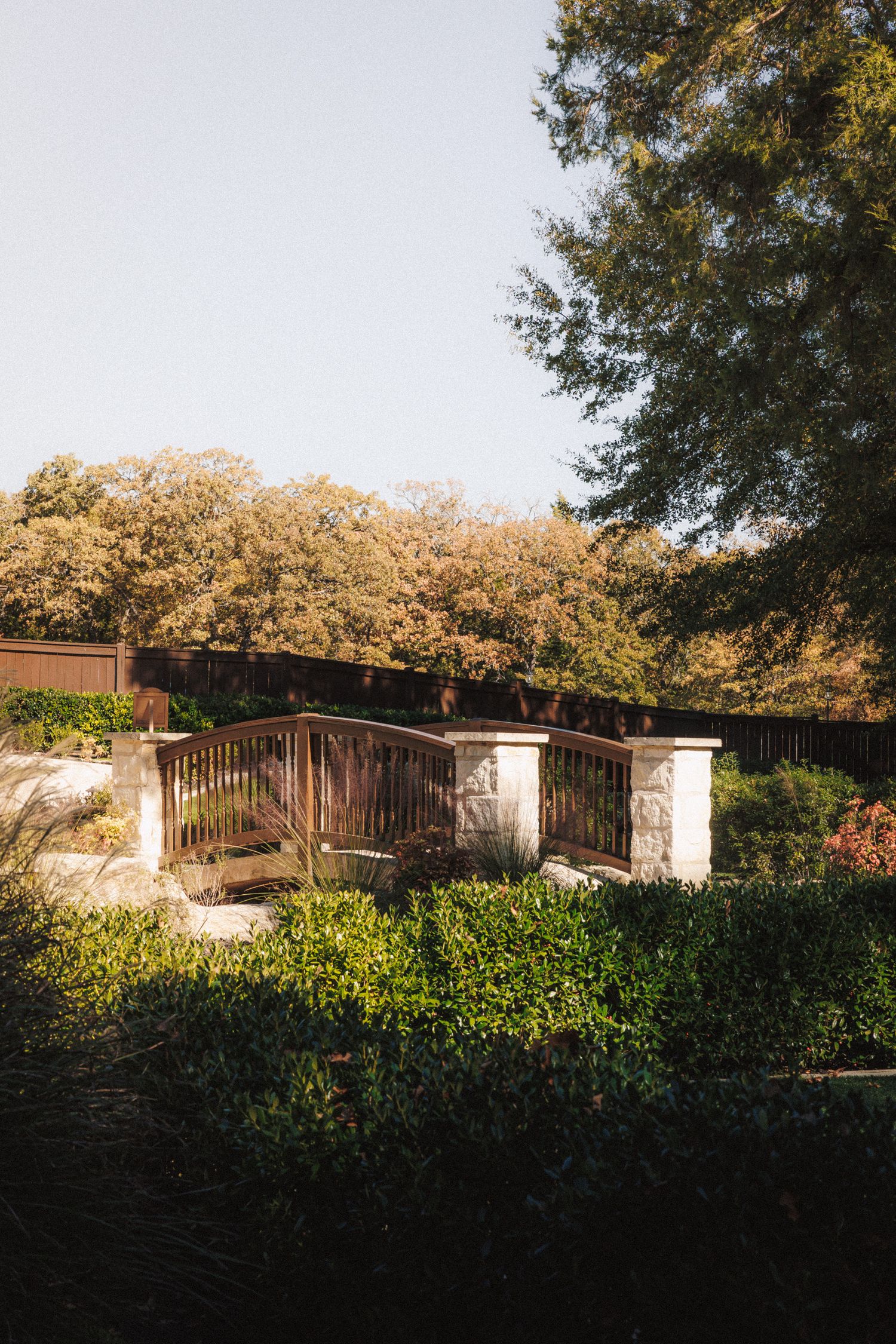 Wooden garden gate with stone pillars surrounded by lush green hedges and tall trees.