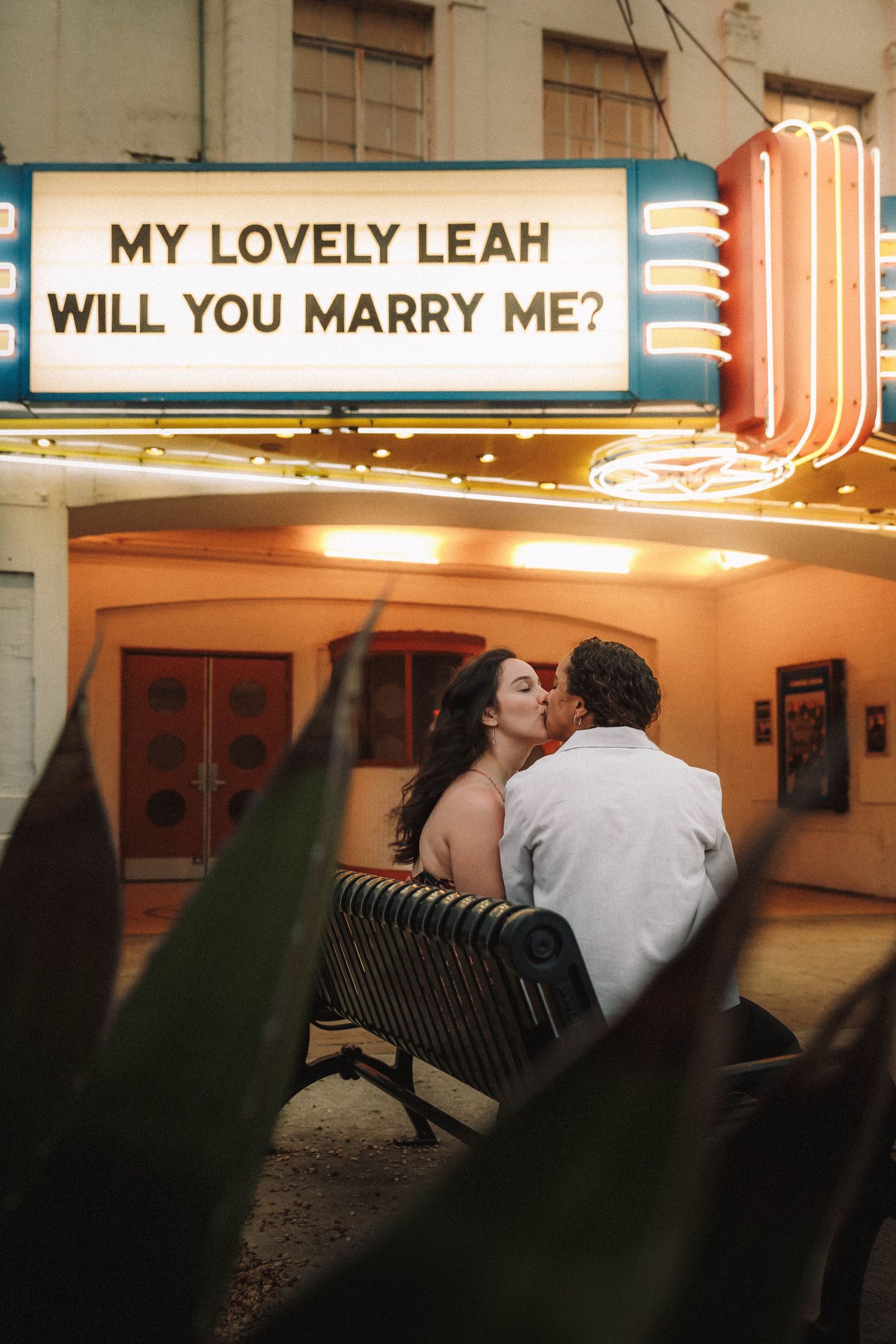 Newly engaged couple kisses on bench under lit marquee proposal sign at night.