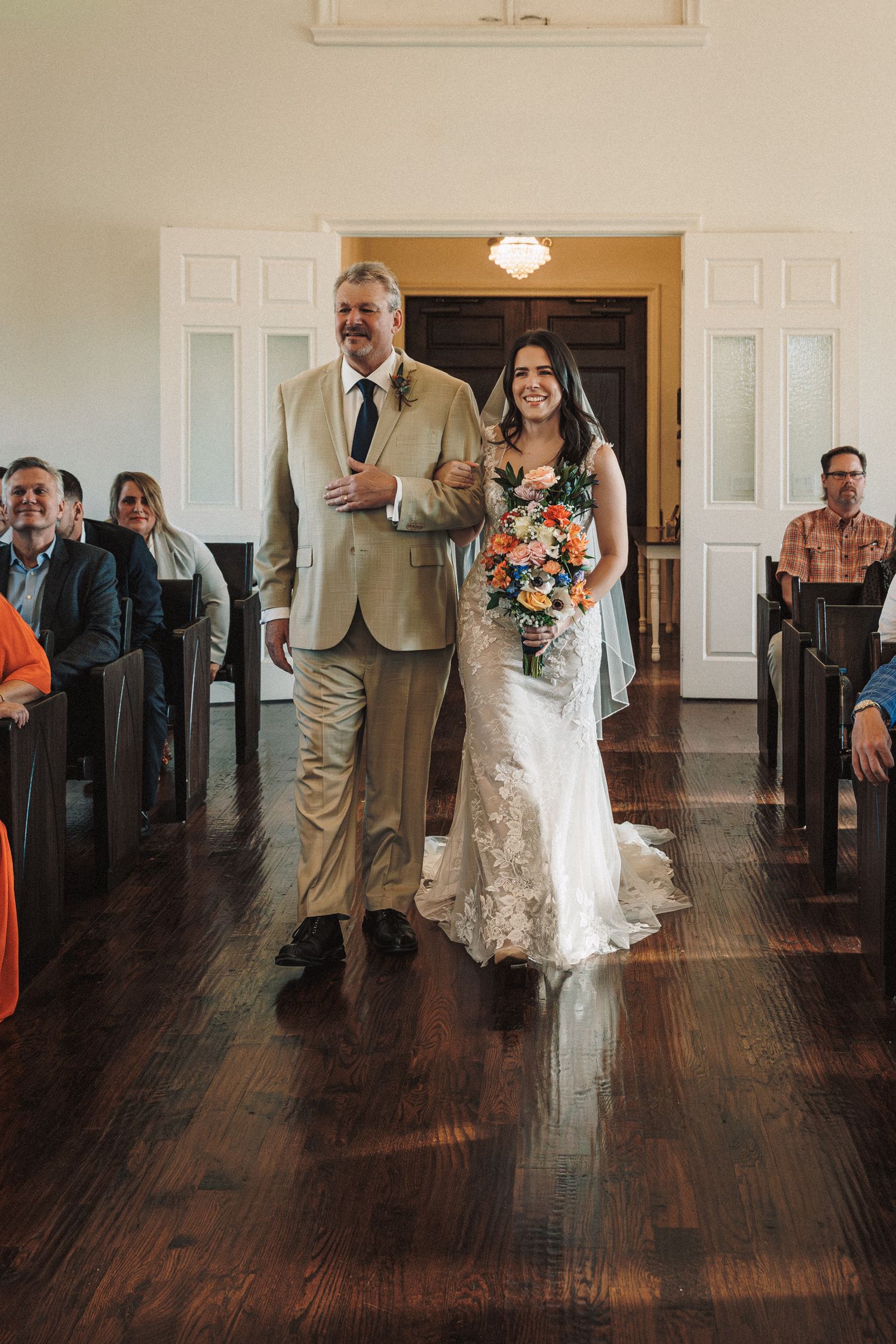 Bride in lace gown walks down aisle with father in tan suit, holding colorful bouquet in elegant wood-floored chapel.