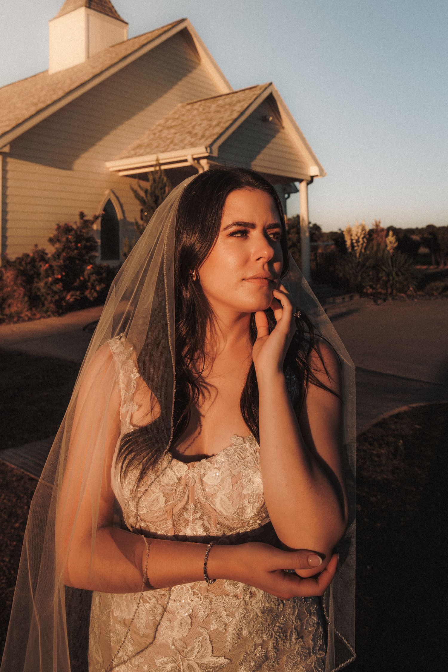 Bride in lace gown and veil poses outside a white chapel at golden hour.