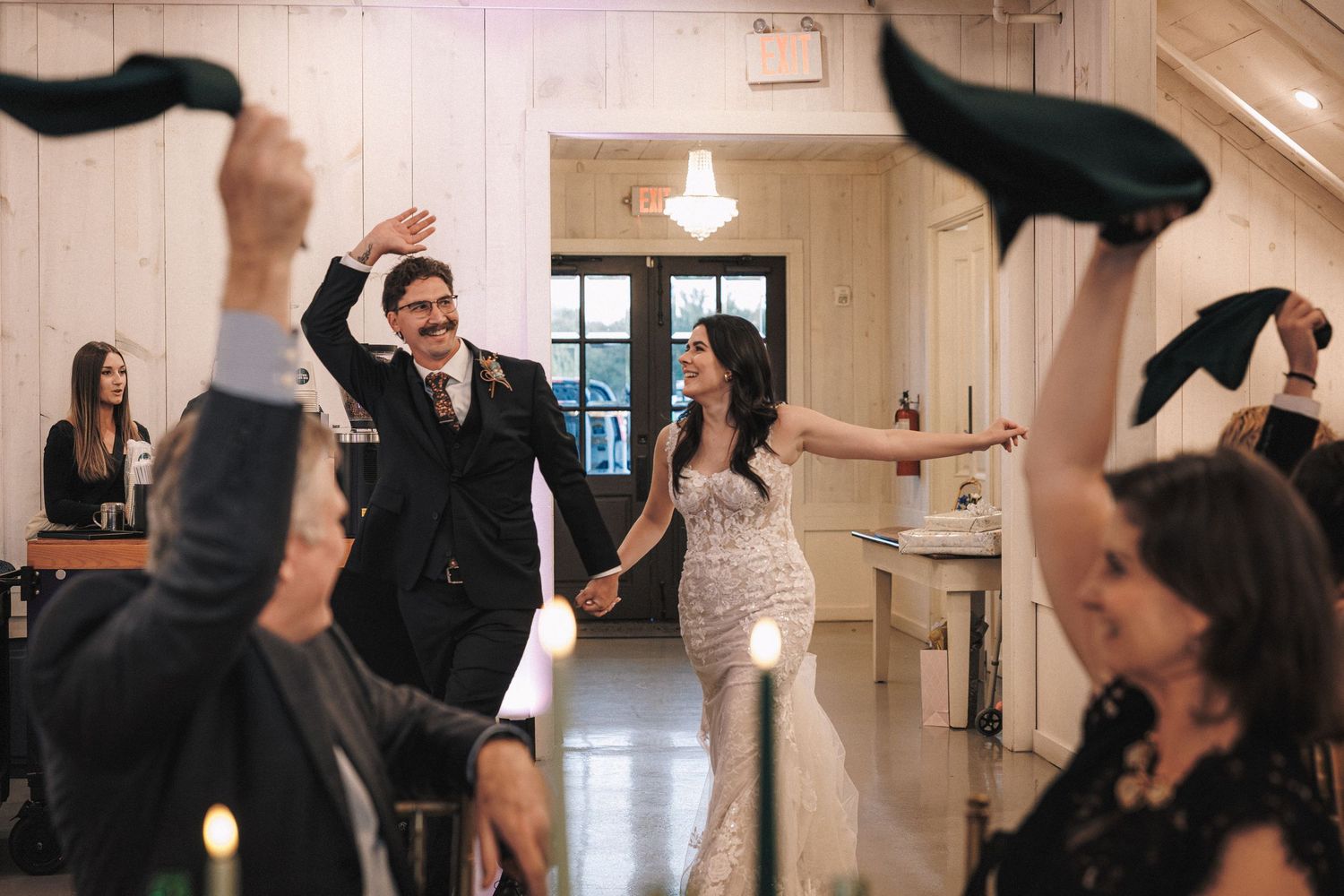 Newlyweds make grand entrance as guests wave hats in celebration inside a white barn wedding venue.