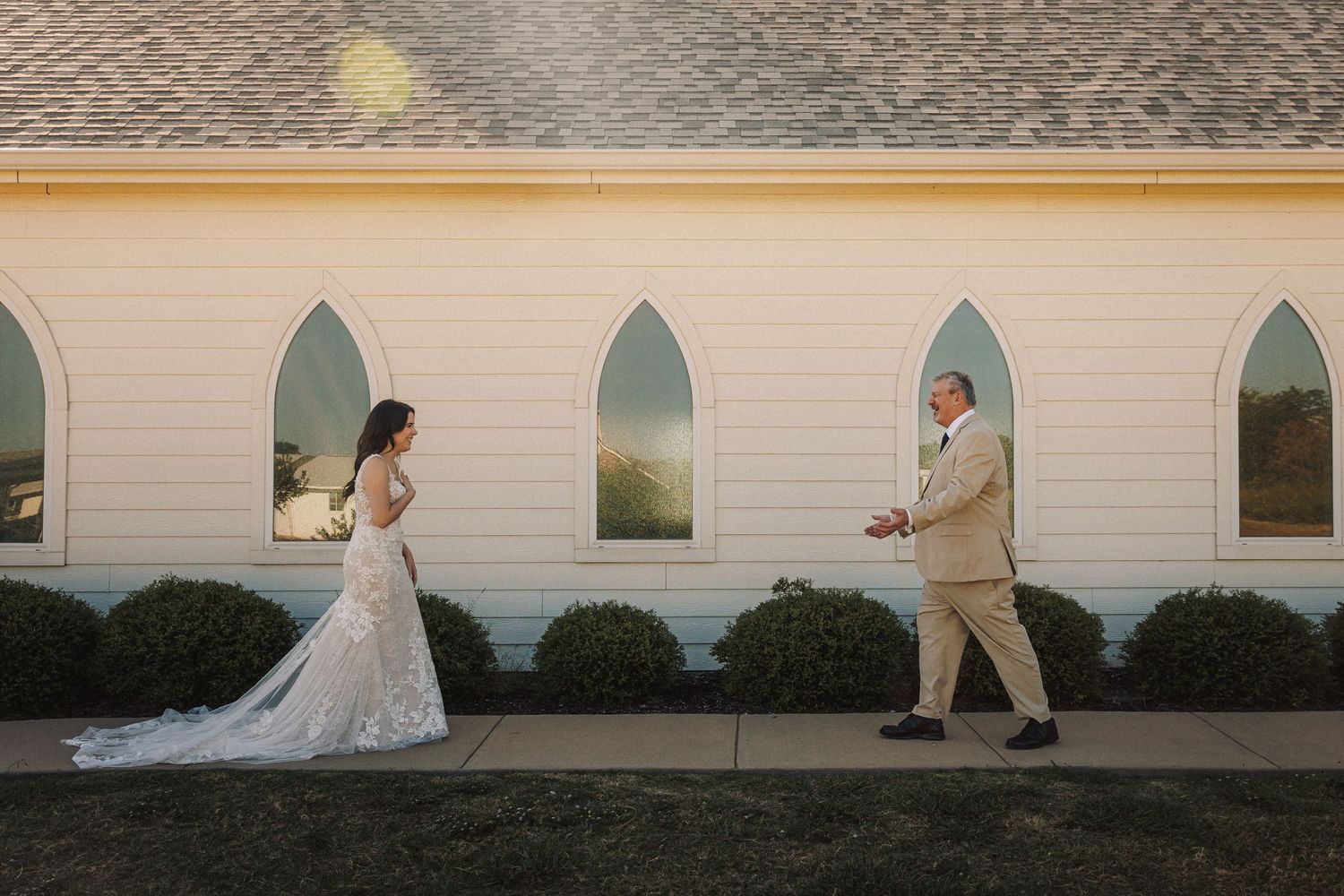 Bride in lace gown and father sharing first look outside white chapel at sunset.
