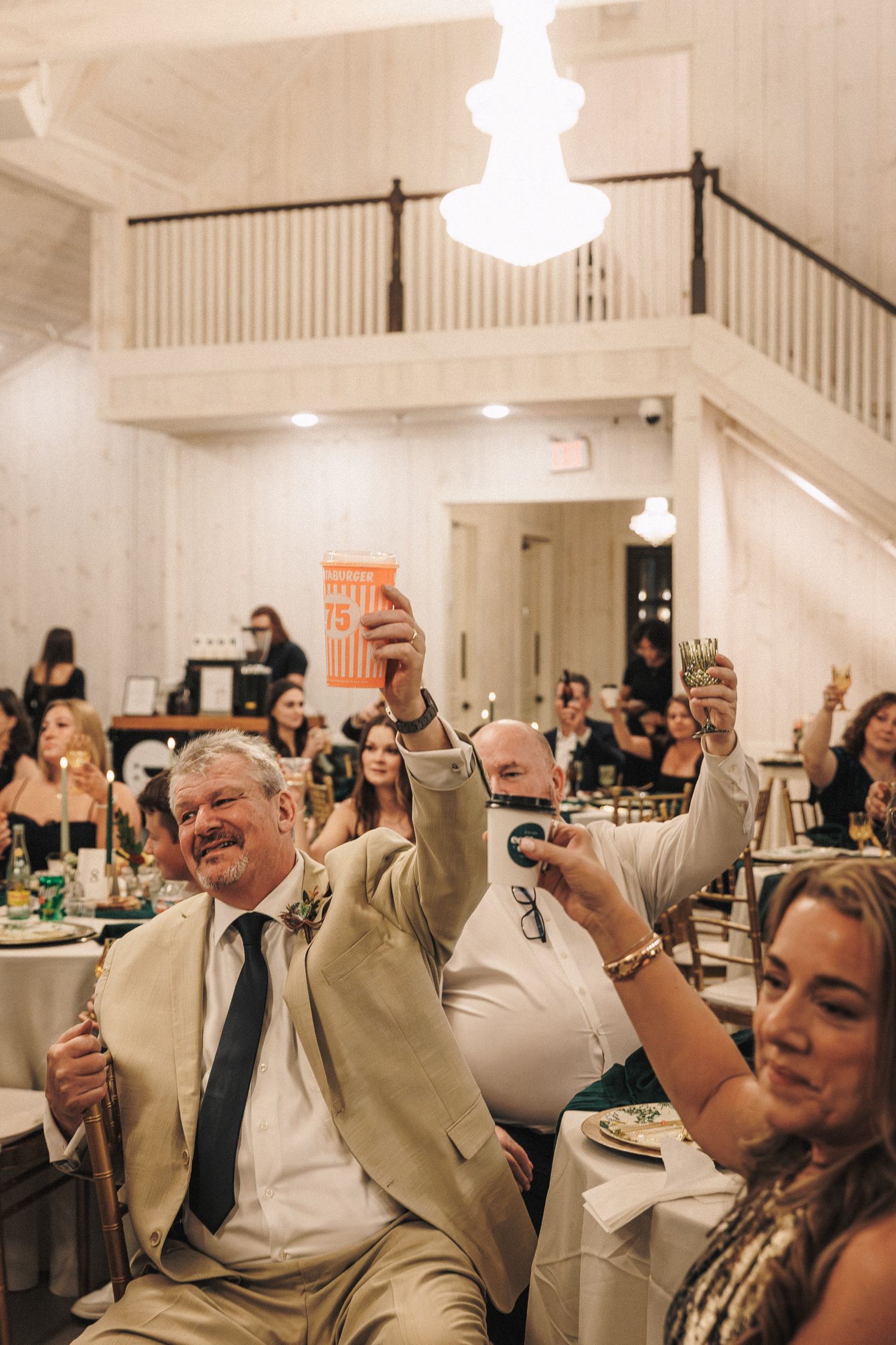 Wedding guests raise glasses in a toast during reception in a warmly lit rustic white barn venue.