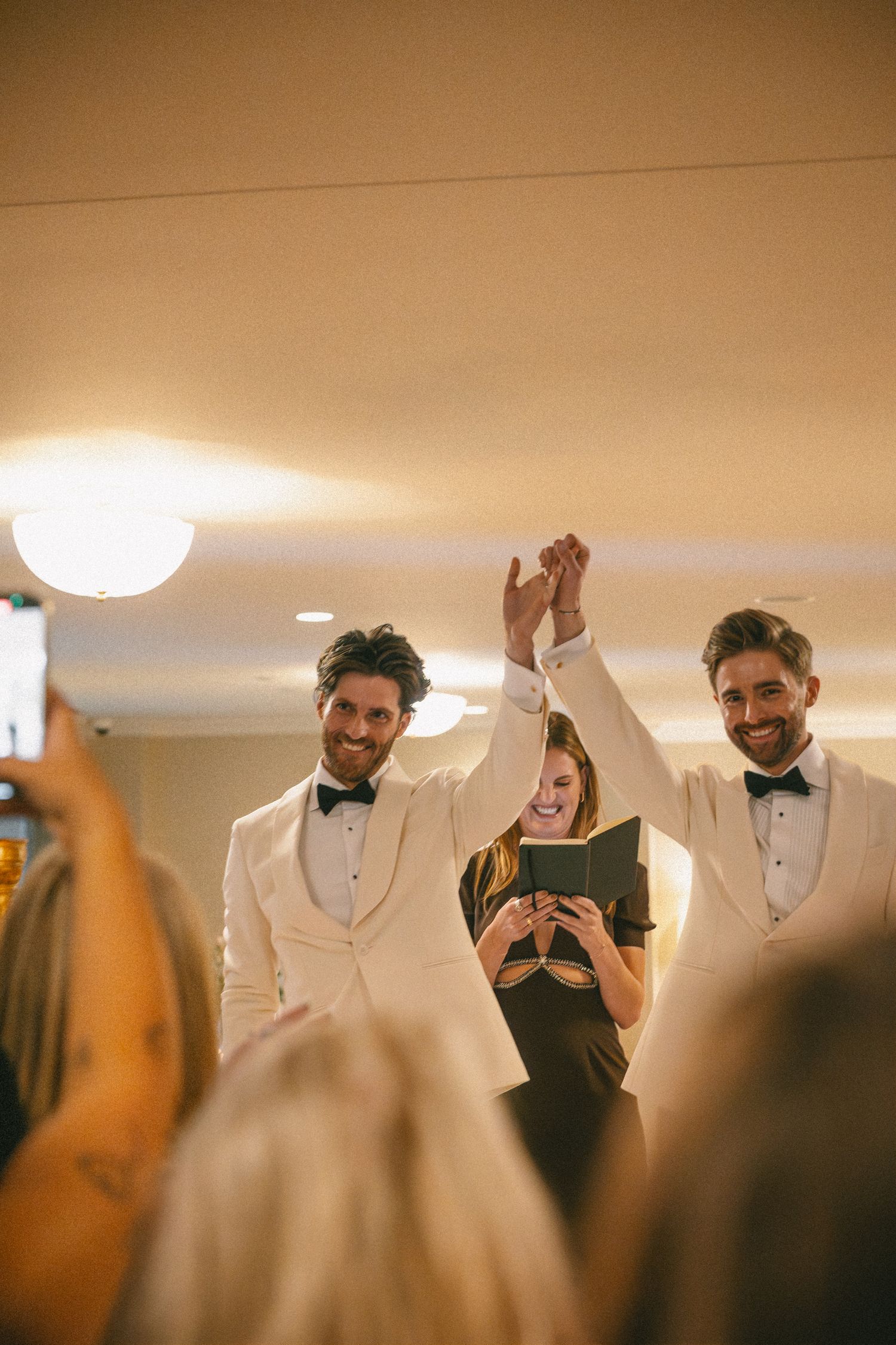 Two grooms in white tuxedos raise hands with officiant at same-sex wedding ceremony.