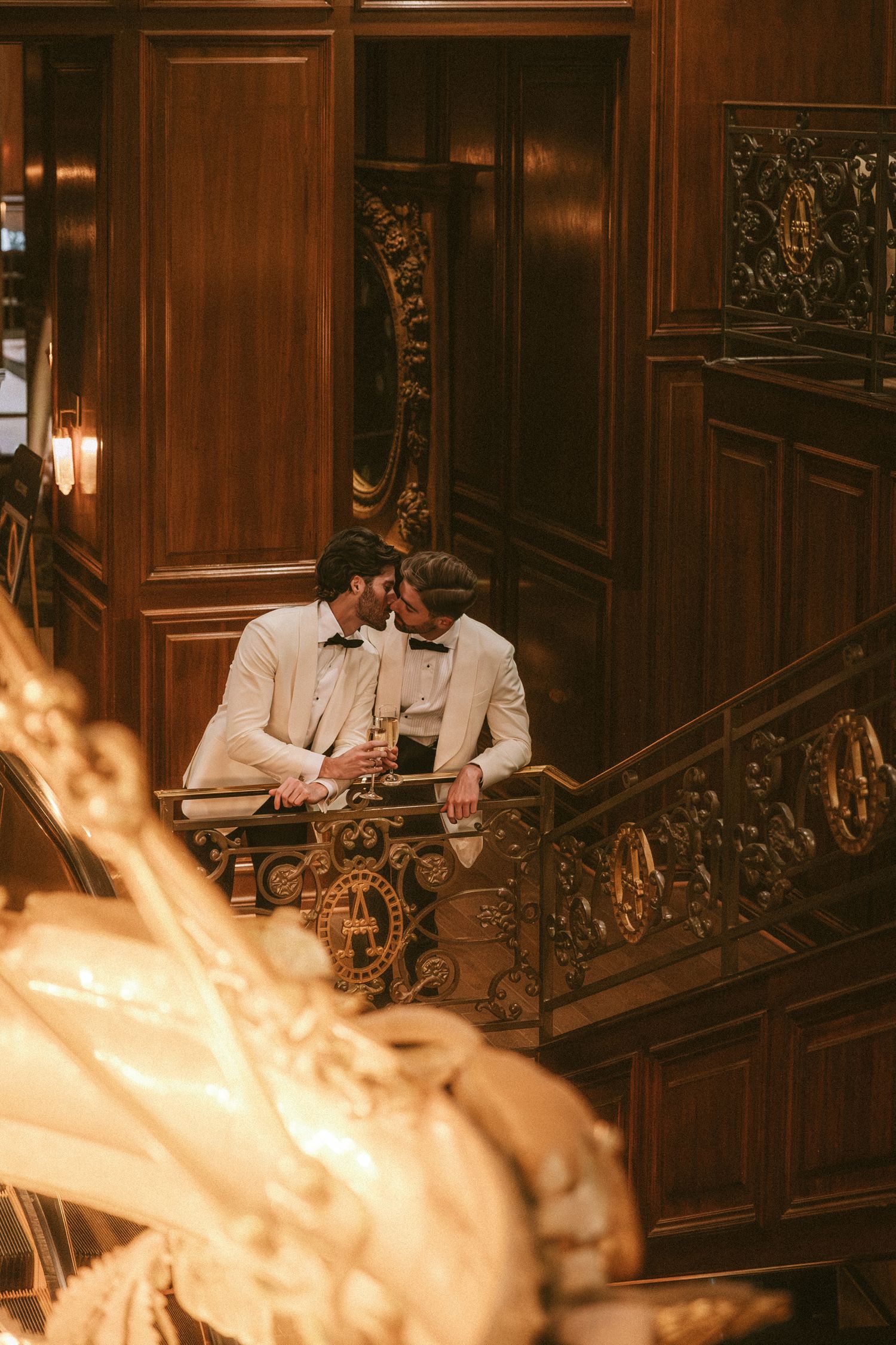 Two grooms sharing intimate moment on balcony of ornate wood-paneled venue.