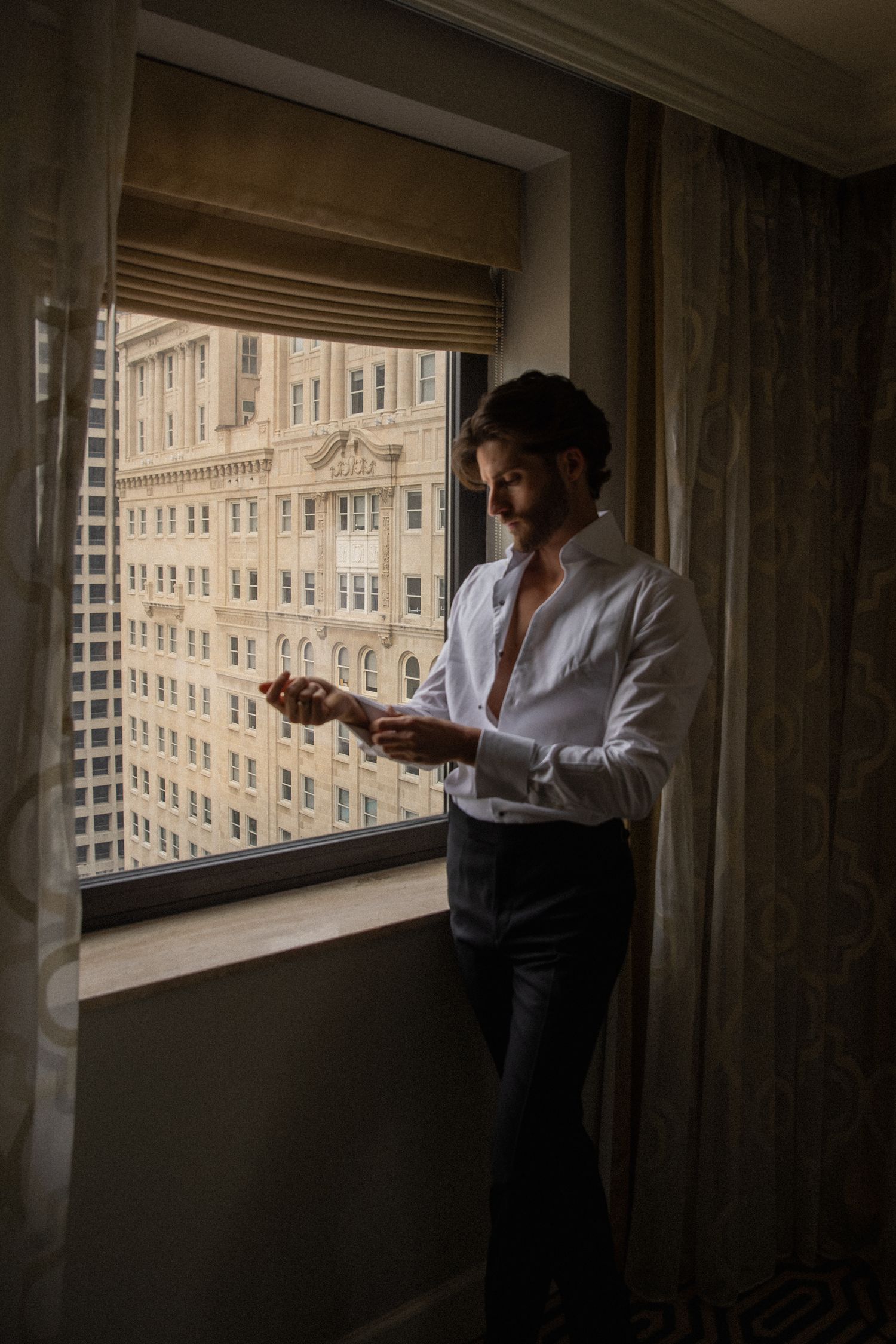 Groom in white dress shirt adjusting cufflinks by hotel window with city building view.