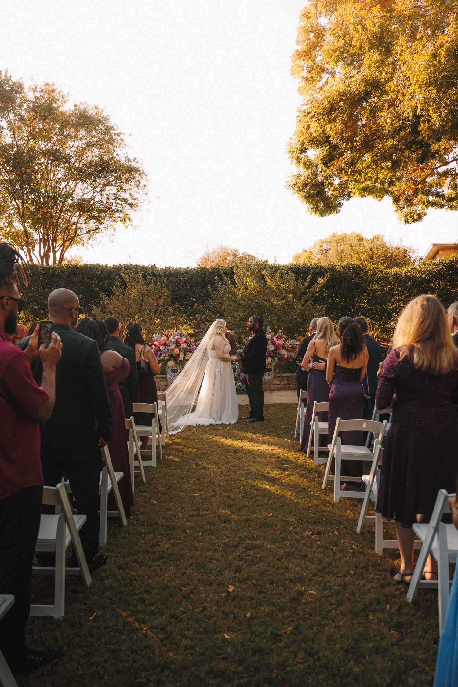 Outdoor wedding ceremony at sunset with bride and groom at altar lined with white chairs and guests.
