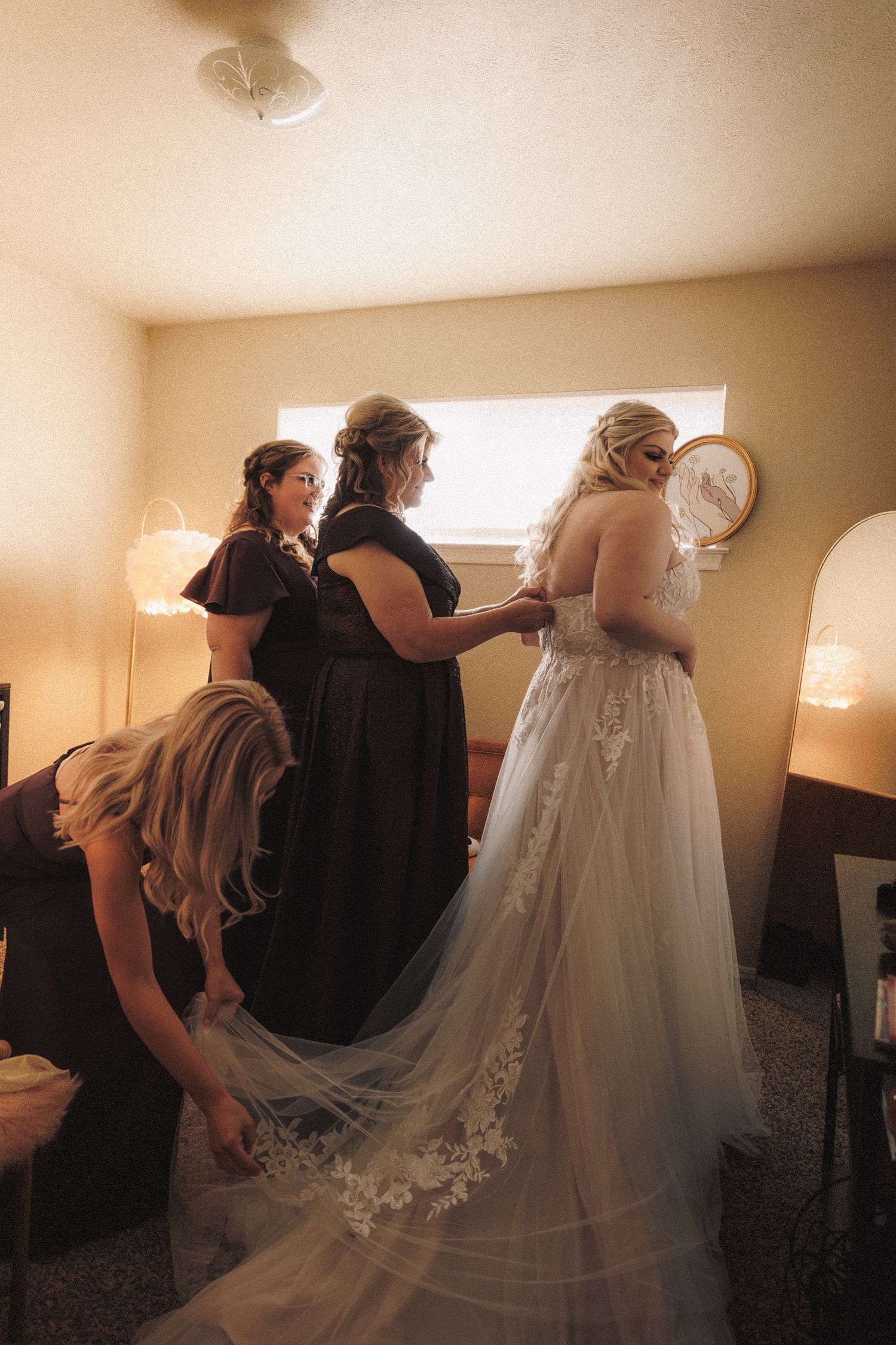 Bridesmaids in dark gowns helping bride into lace wedding dress with cathedral train in warm-lit room.