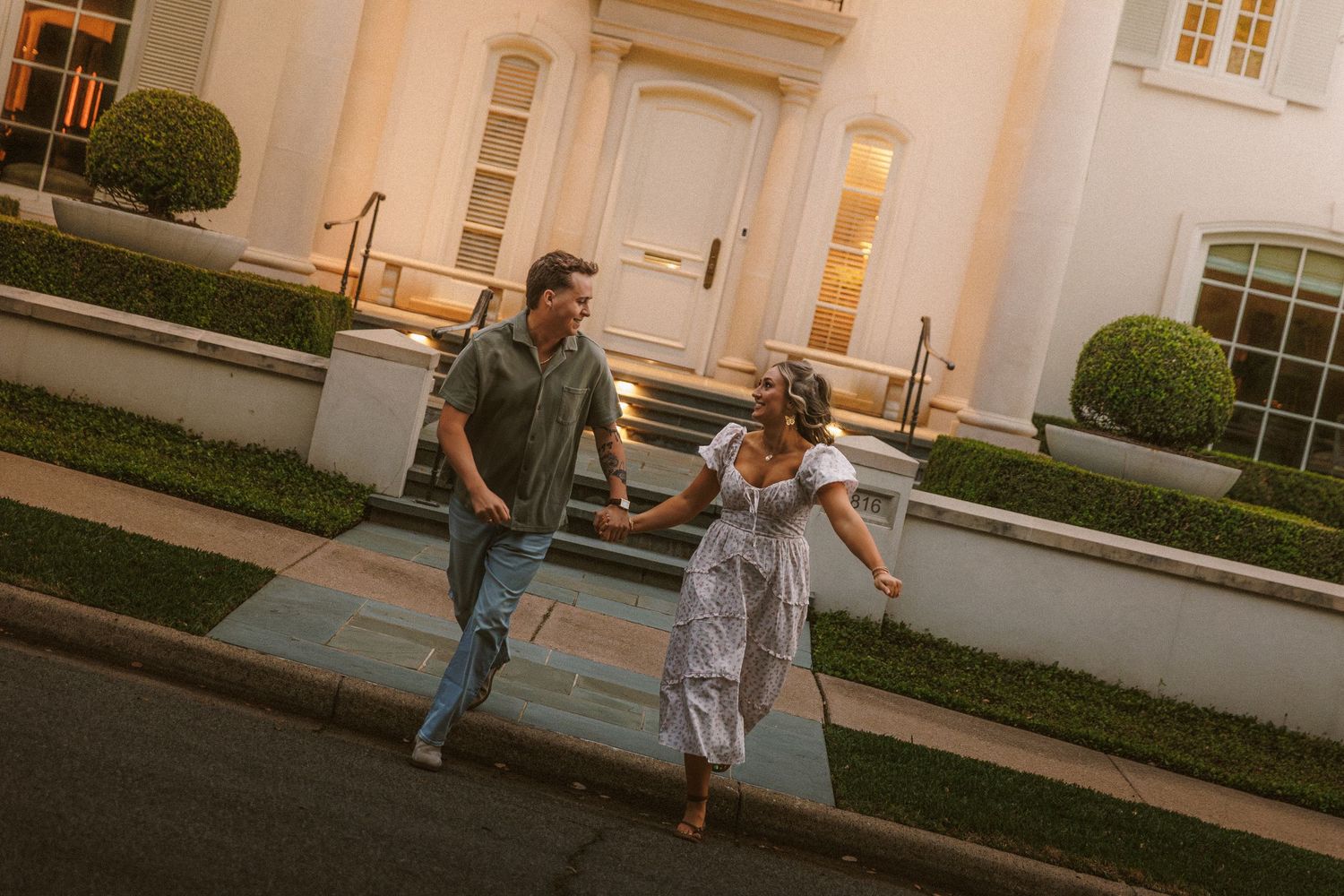 Couple holding hands and running down steps in front of an elegant white mansion at golden hour.