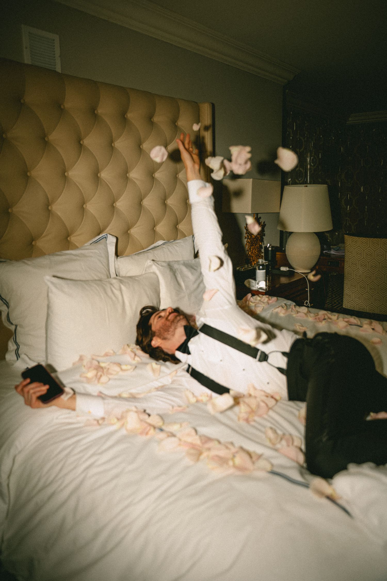 Groom in white shirt and suspenders tosses rose petals on decorated hotel suite bed.