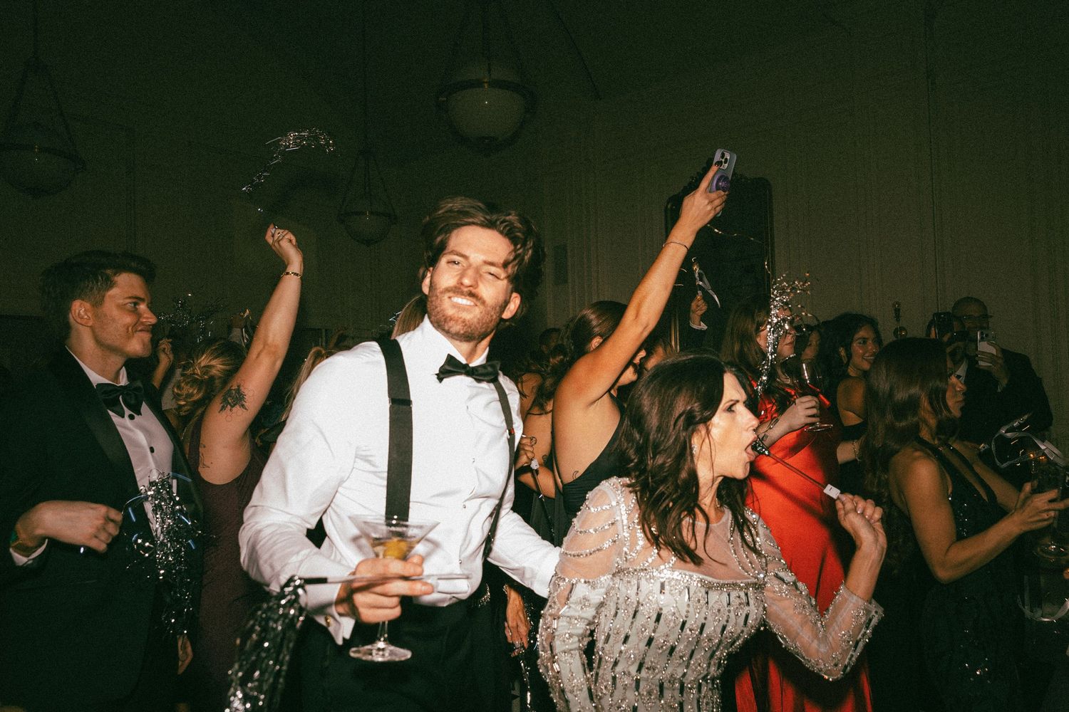 Groom in suspenders dancing and raising a glass with guests at a lively wedding reception.