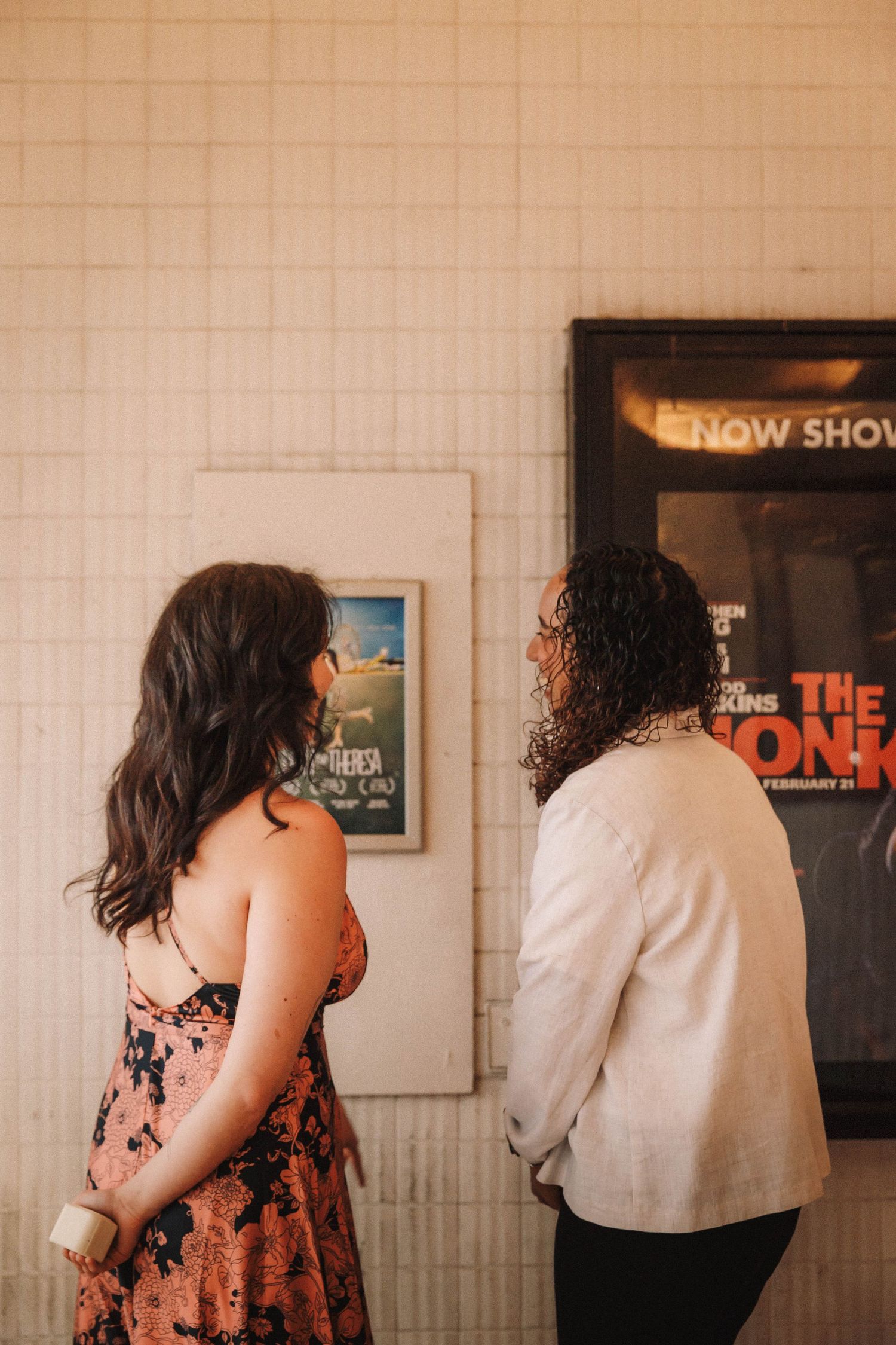 Couple looking at movie posters on a tiled wall inside a vintage cinema theater.