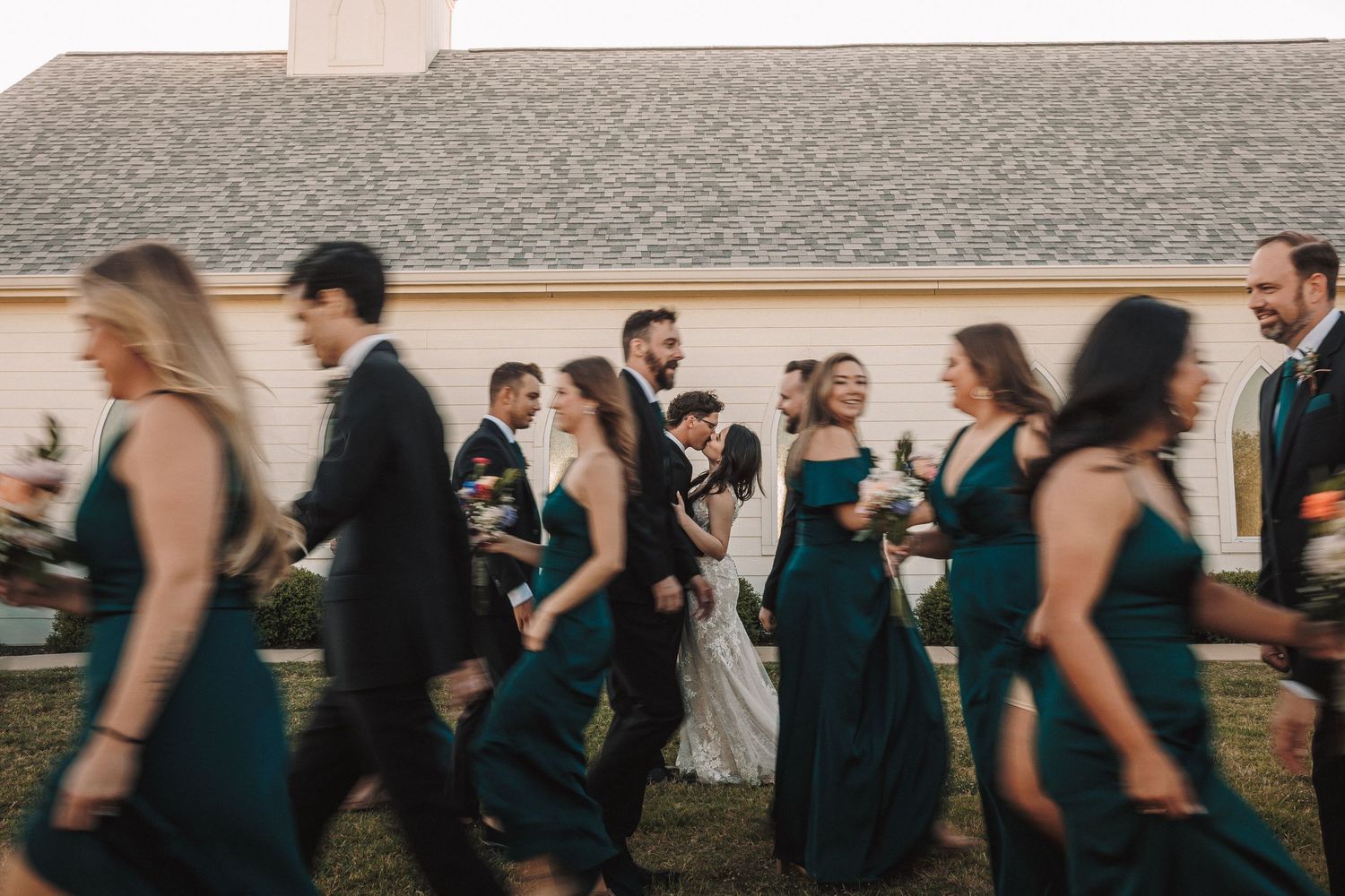 Wedding party walks together in emerald green and black attire outside chapel.